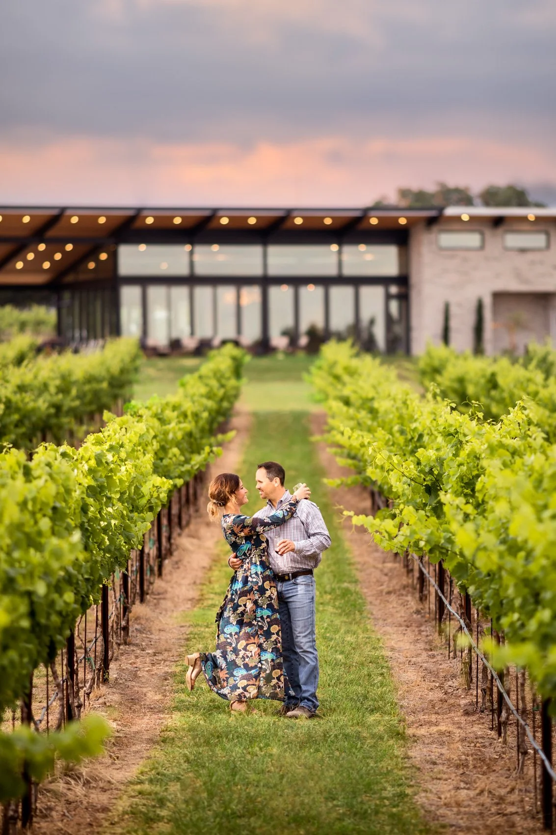 A couple embrace and smile in a vineyard with lush green grapevines, a modern building with large windows and string lights in the background at sunset.