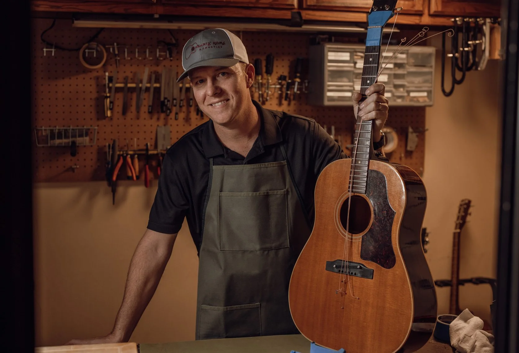 A smiling man in a garage workshop holding an acoustic guitar.