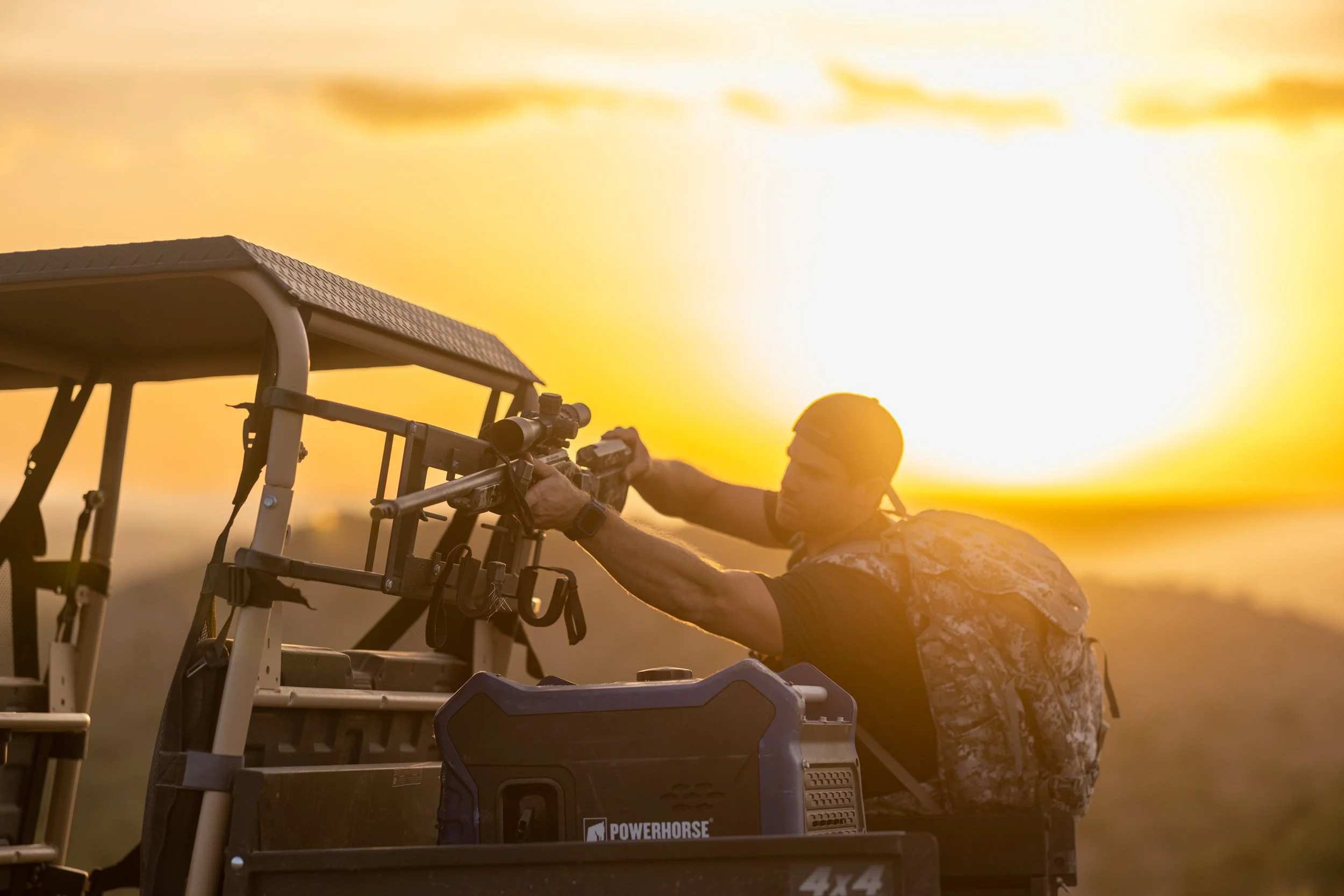 A person wearing camouflage-patterned clothing and an orange hat is aiming a scoped rifle mounted on a hunting vehicle during sunset.