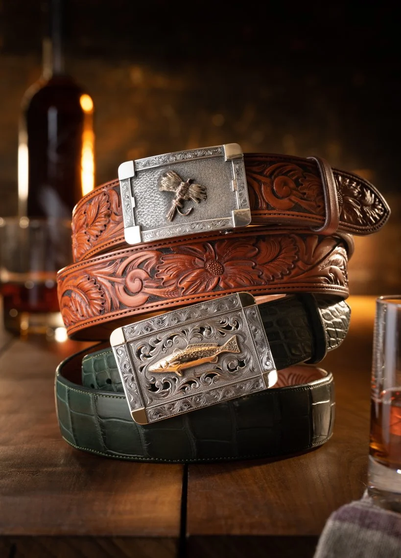 Three leather belts with decorative belt buckles stacked on a wooden table, with a blurred background featuring a bottle and glasses.