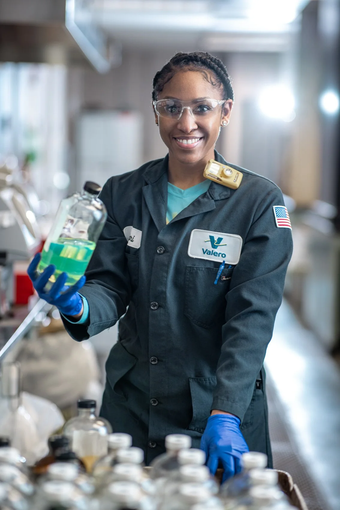 A smiling woman in safety glasses and a Valero uniform holding a beaker with a green liquid in a laboratory or industrial setting.