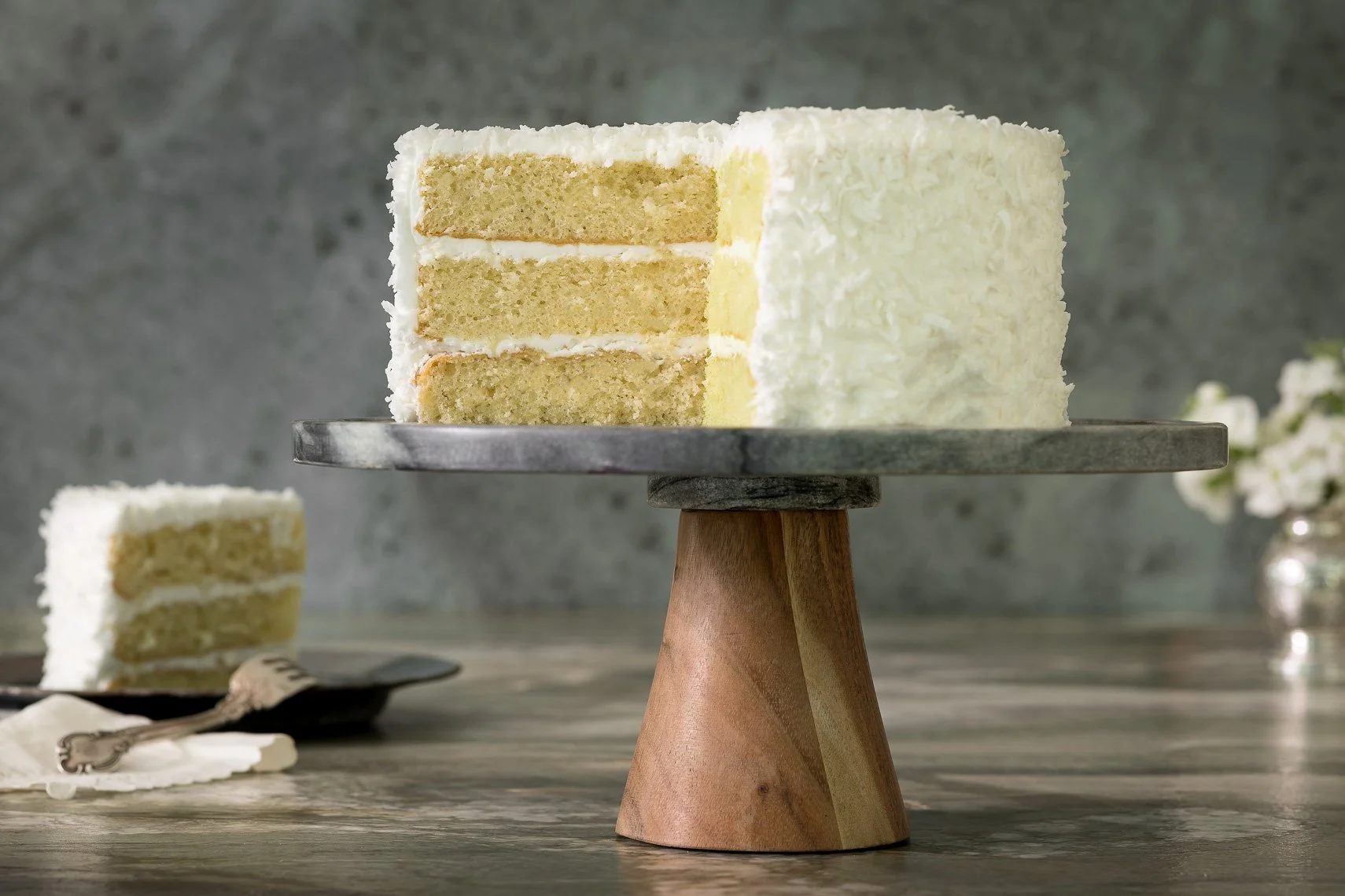 A slice of layered white cake with cream filling on a black plate, with a fork and a small piece of cake on the side, placed on a wooden table.