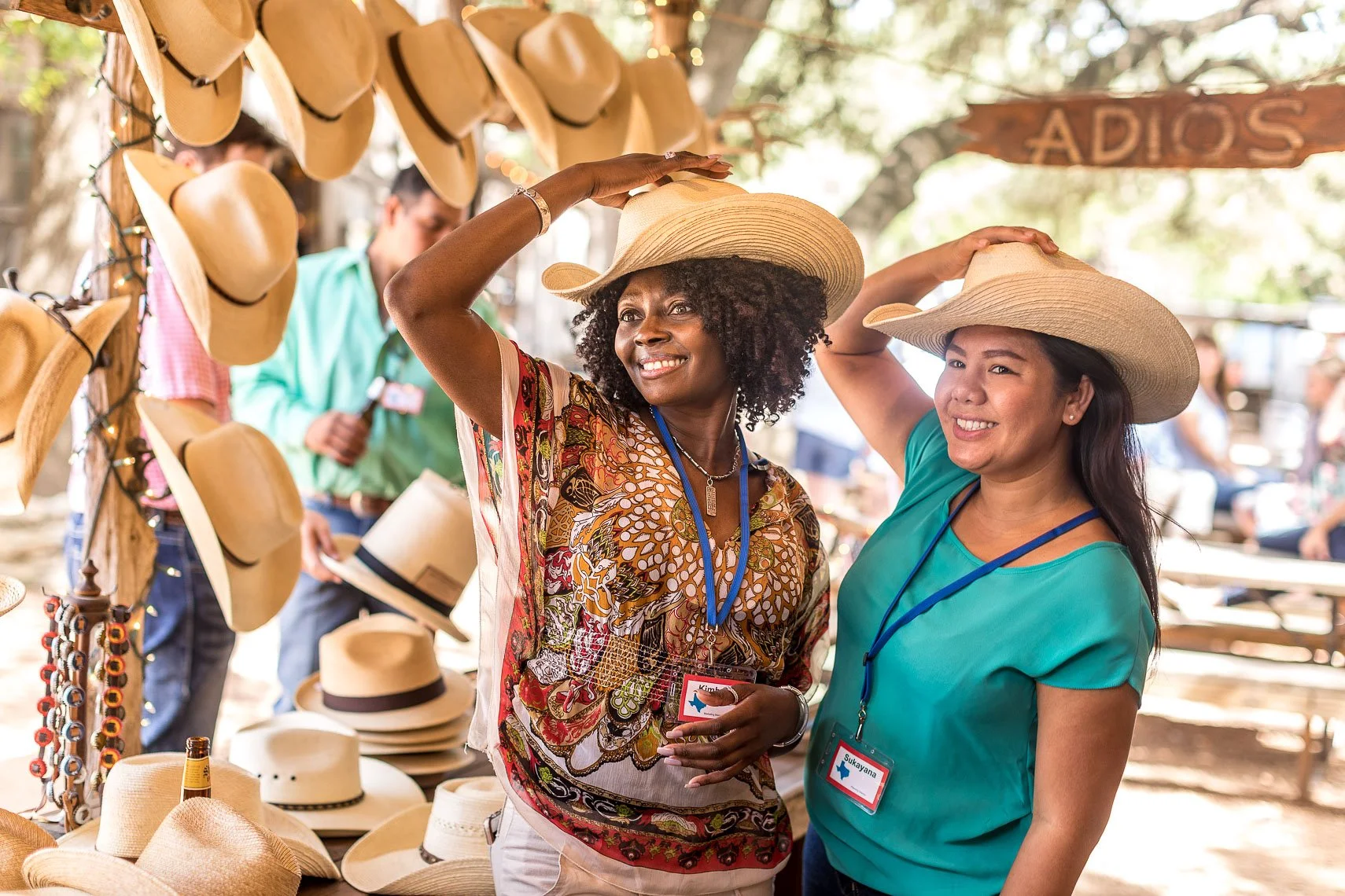 Two women try on straw hats at an outdoor market stand, smiling and wearing name tags, with hats and accessories displayed on the stand behind them.