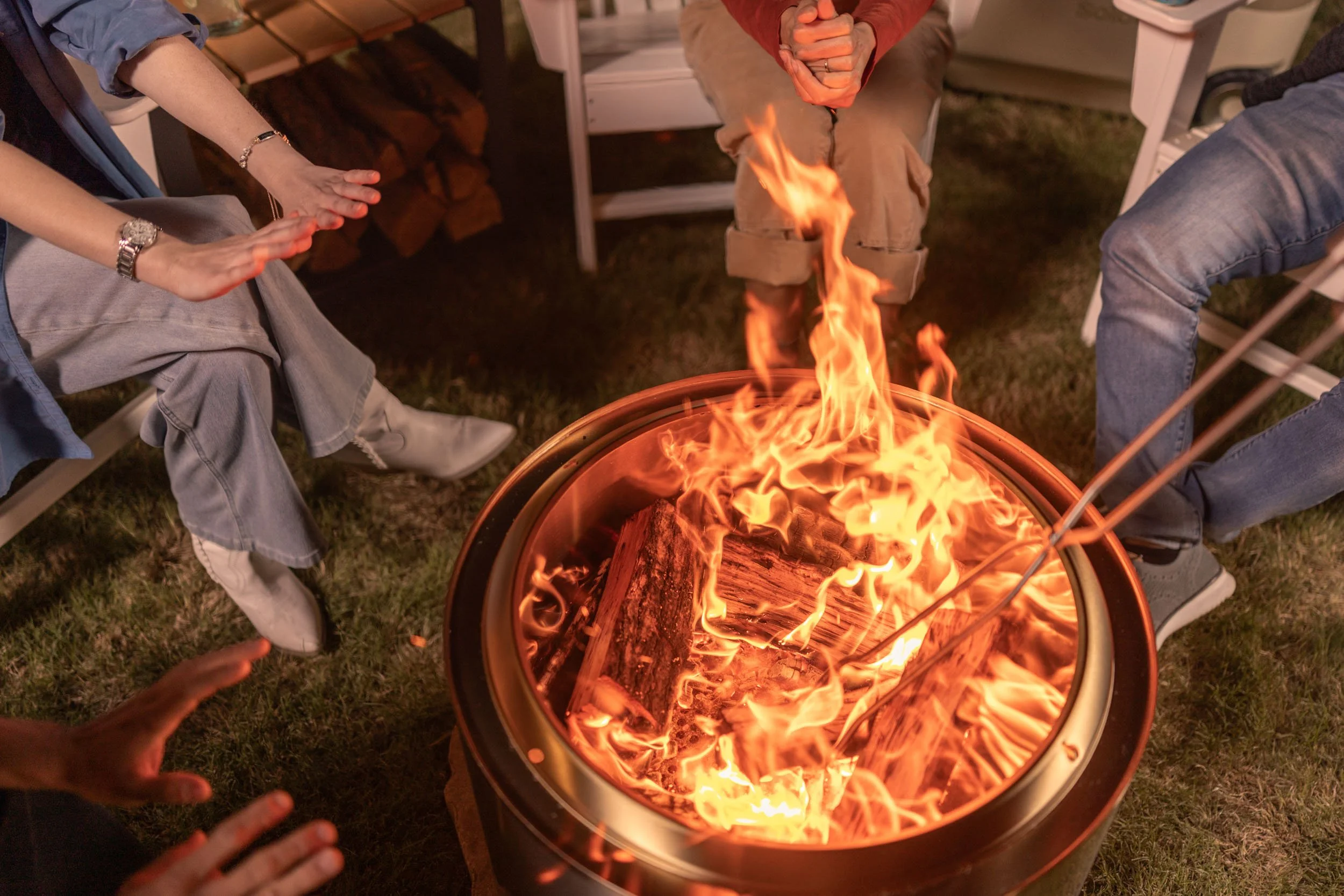People sitting around a fire pit, some with their hands extended towards the flames, outdoors at night.