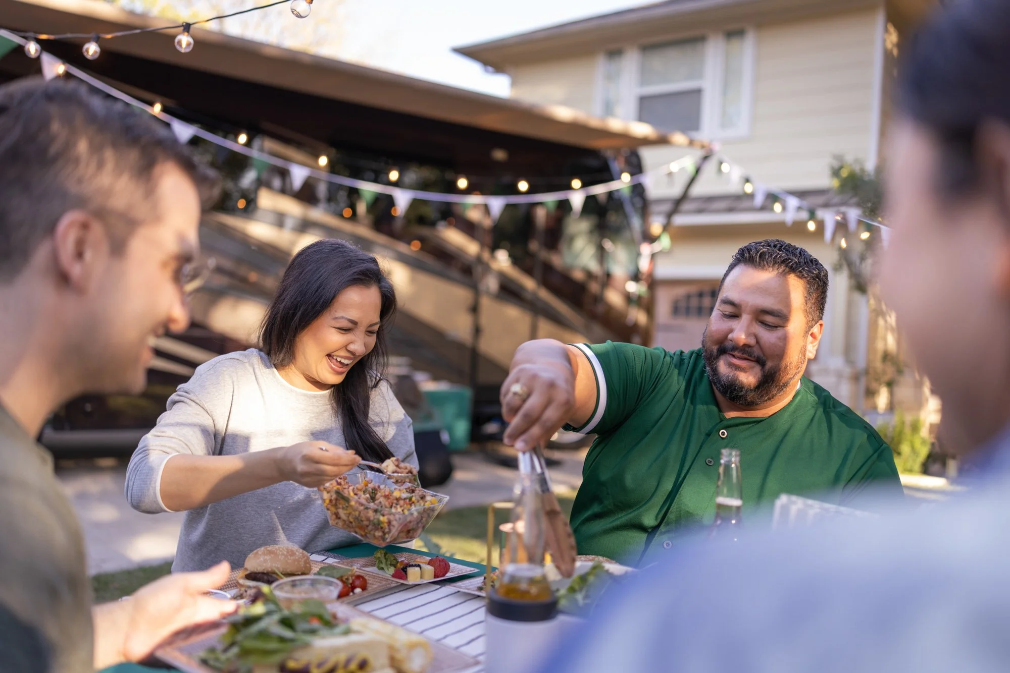 People gather outdoors around a table with food at a backyard barbecue, decorated with string lights and bunting.