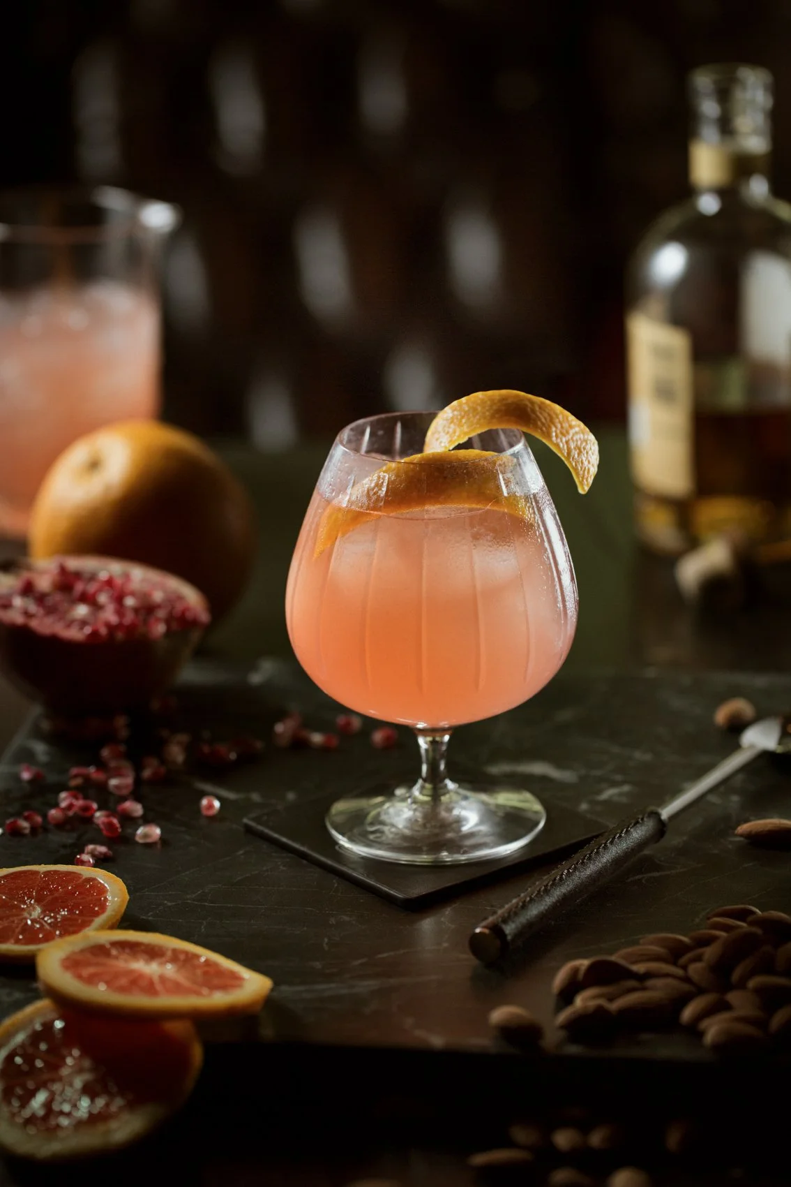 A pink cocktail with a lemon twist garnish in a stemmed glass, surrounded by citrus fruits, pomegranate seeds, and nuts on a dark table.