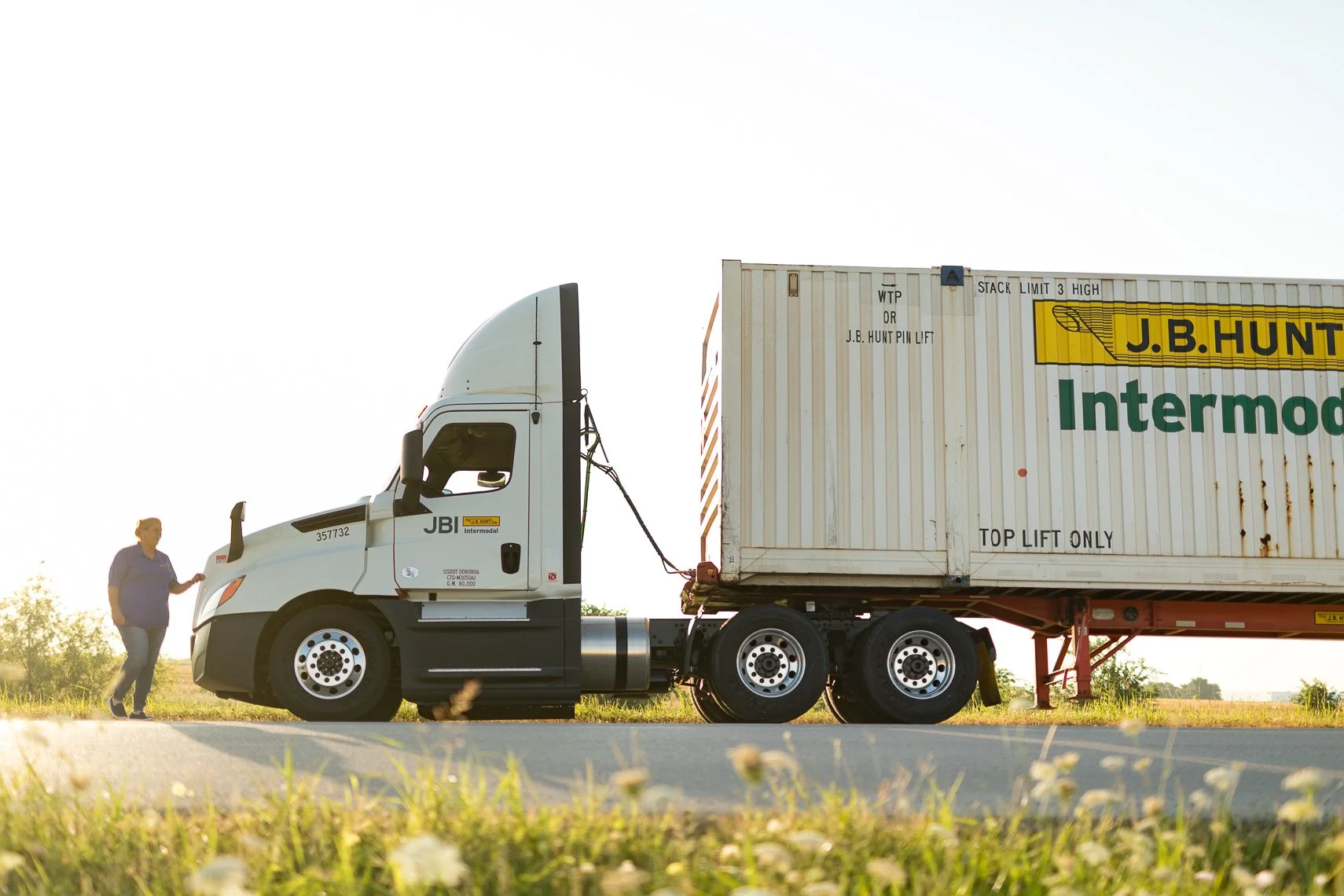 A person standing outside next to a white semi-truck with a large trailer. The trailer has a yellow and green logo that reads "J.B. Hunt" and the word "Intermodal." The person appears to be inspecting or waiting, with grassy land and trees in the bac