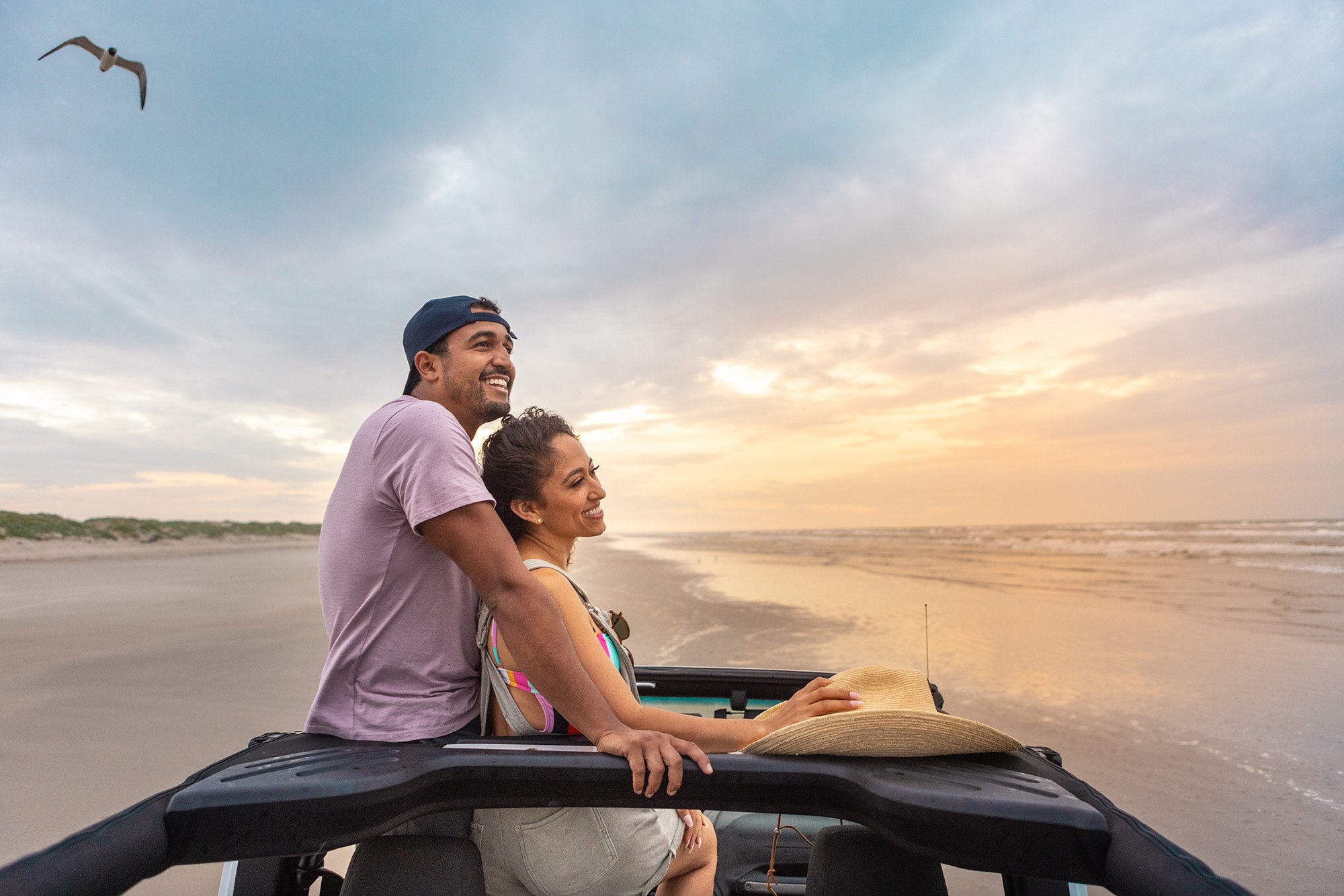 A smiling man and woman ride in an open vehicle along the beach at sunset, with seagulls flying overhead.