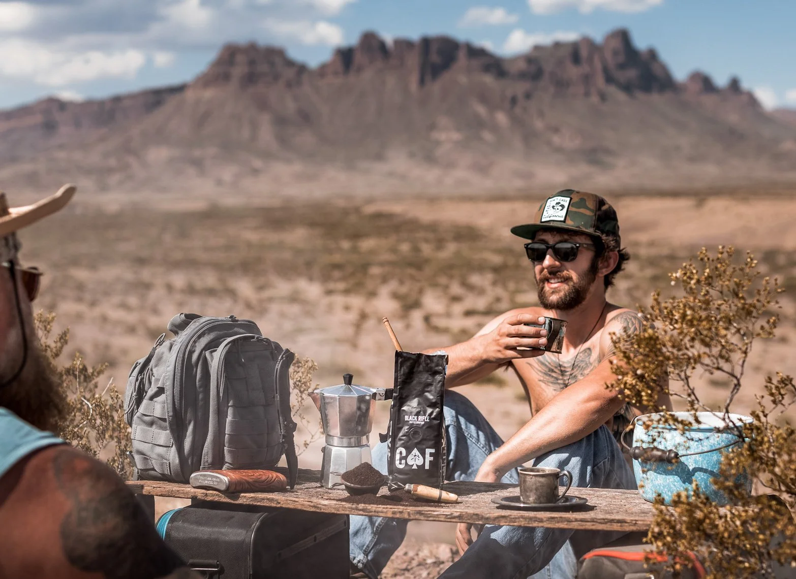 A man sitting at a wooden table outdoors in a desert landscape, wearing sunglasses, a camo hat, and no shirt, holding a cup, with a backpack, a moka pot, a bag of coffee, and other camping items on the table, with mountains in the background.