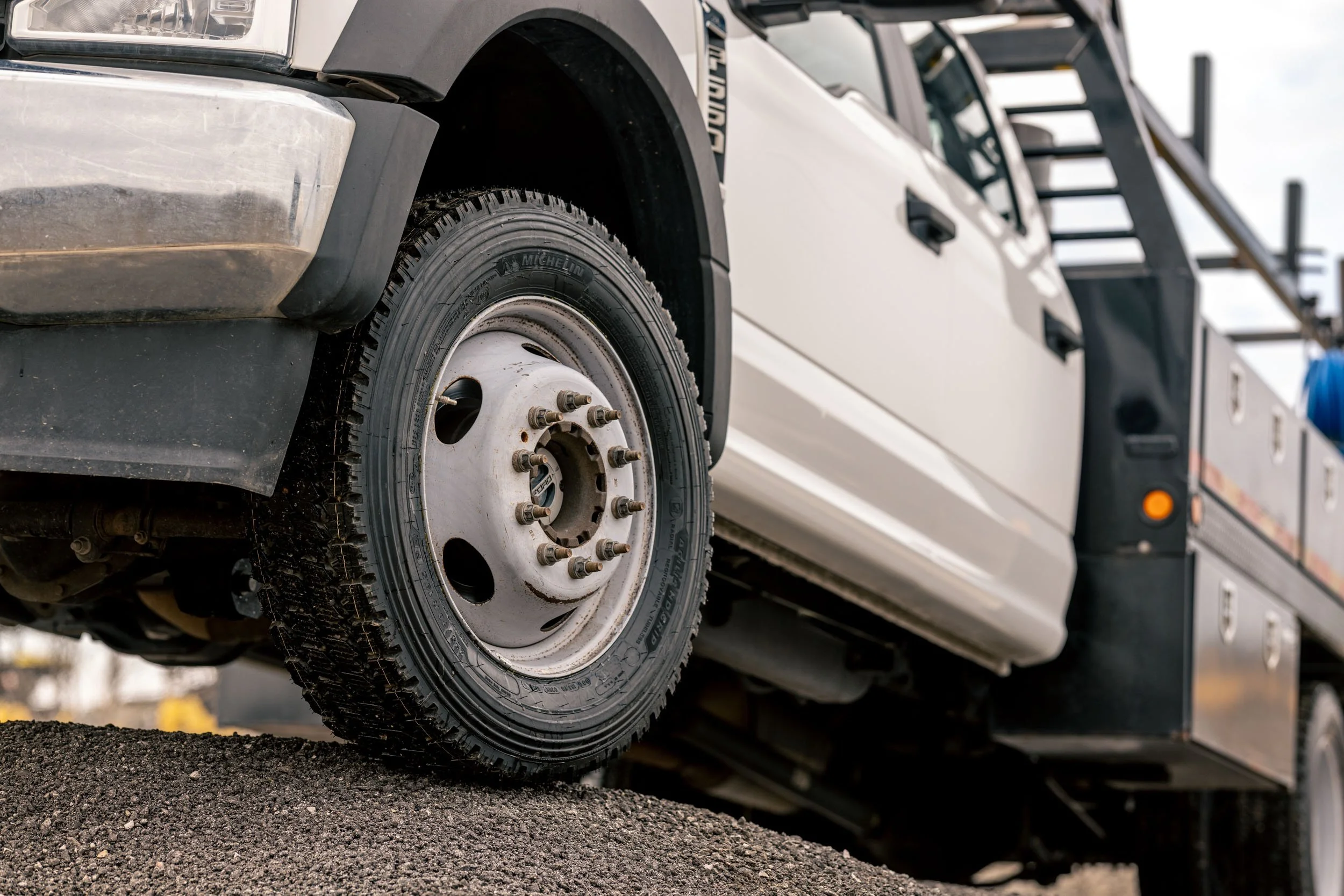 Close-up of a truck tire mounted on a metal rim, with the truck parked on a gravel surface and a ladder on the side of the truck in the background.