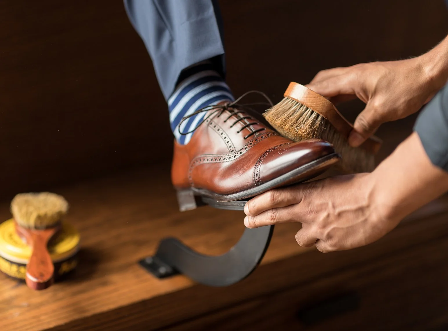 Person polishing a brown leather dress shoe with a horsehair brush, wearing blue pants and striped socks, on a wooden surface.