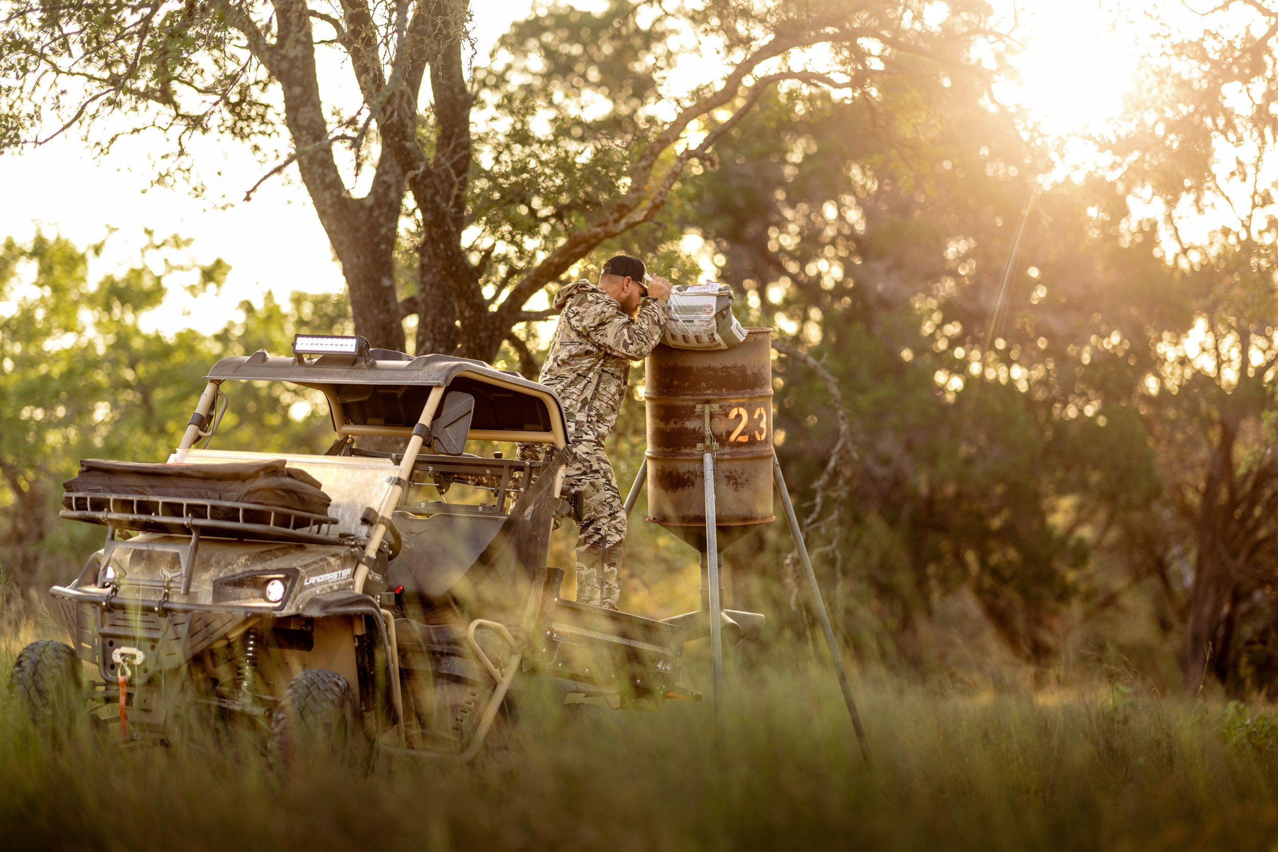 A person in camouflage clothing stands outdoors near a rusty barrel labeled with the number 23, examining or using a rectangular object on top of the barrel. There is a four-wheeler vehicle parked nearby, and the scene is set in a wooded area with su