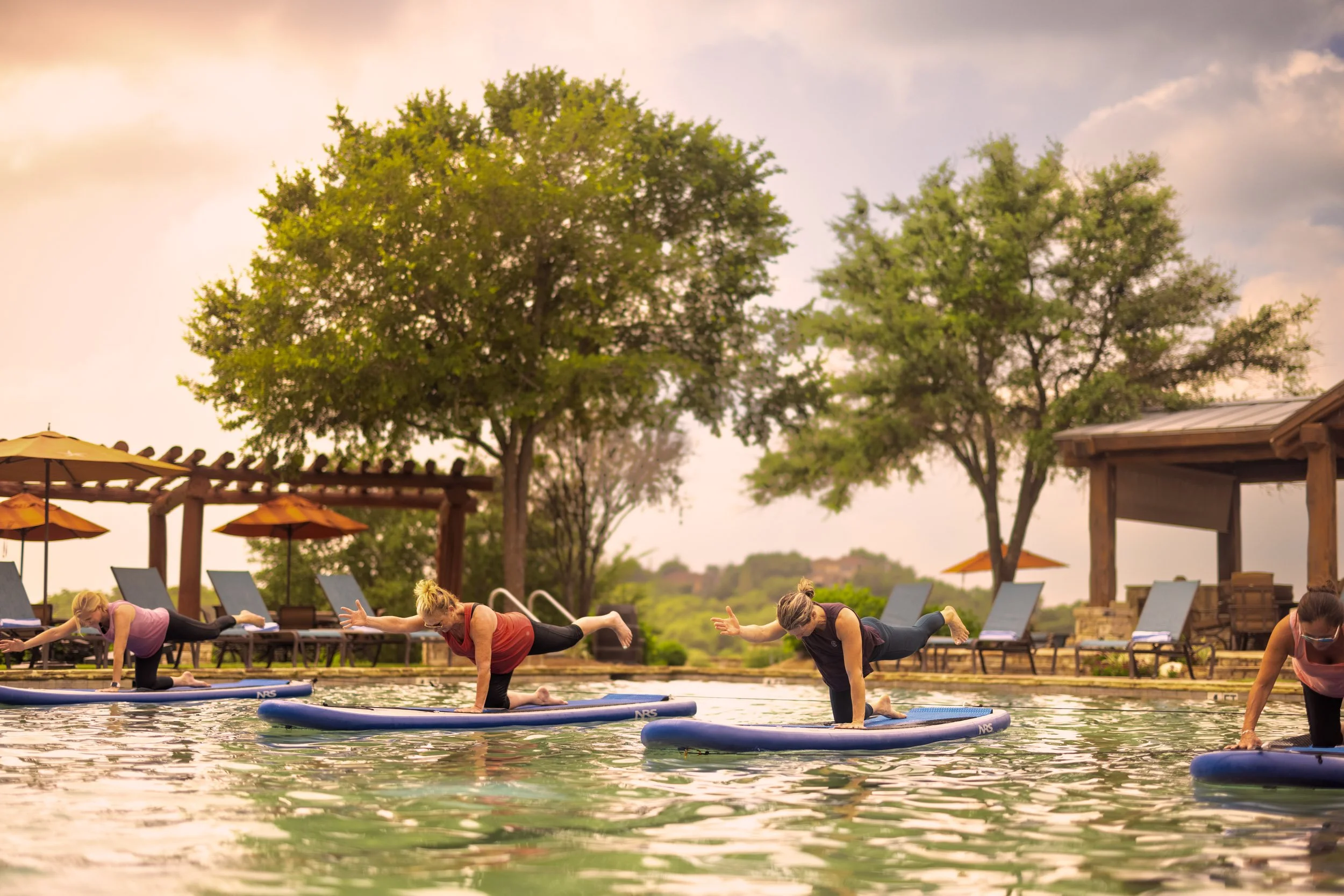 Group of people practicing yoga on paddleboards in a pool outdoors during sunset, with trees and umbrellas in the background.