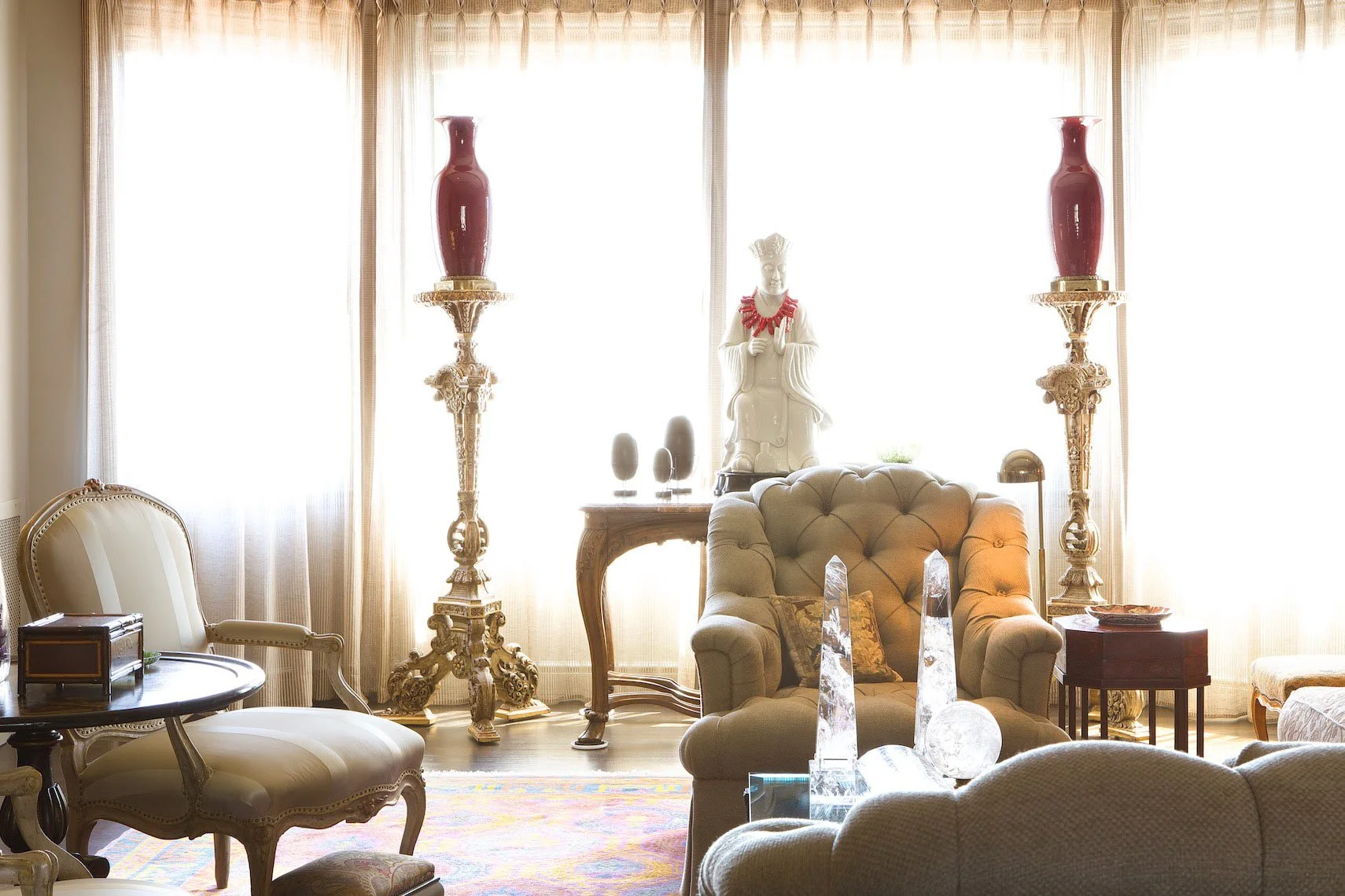 Living room with a large window, two tall red vases on ornate gold stands, a white statue on a table, a cream chaise lounge, a vintage armchair, and various decorative objects.