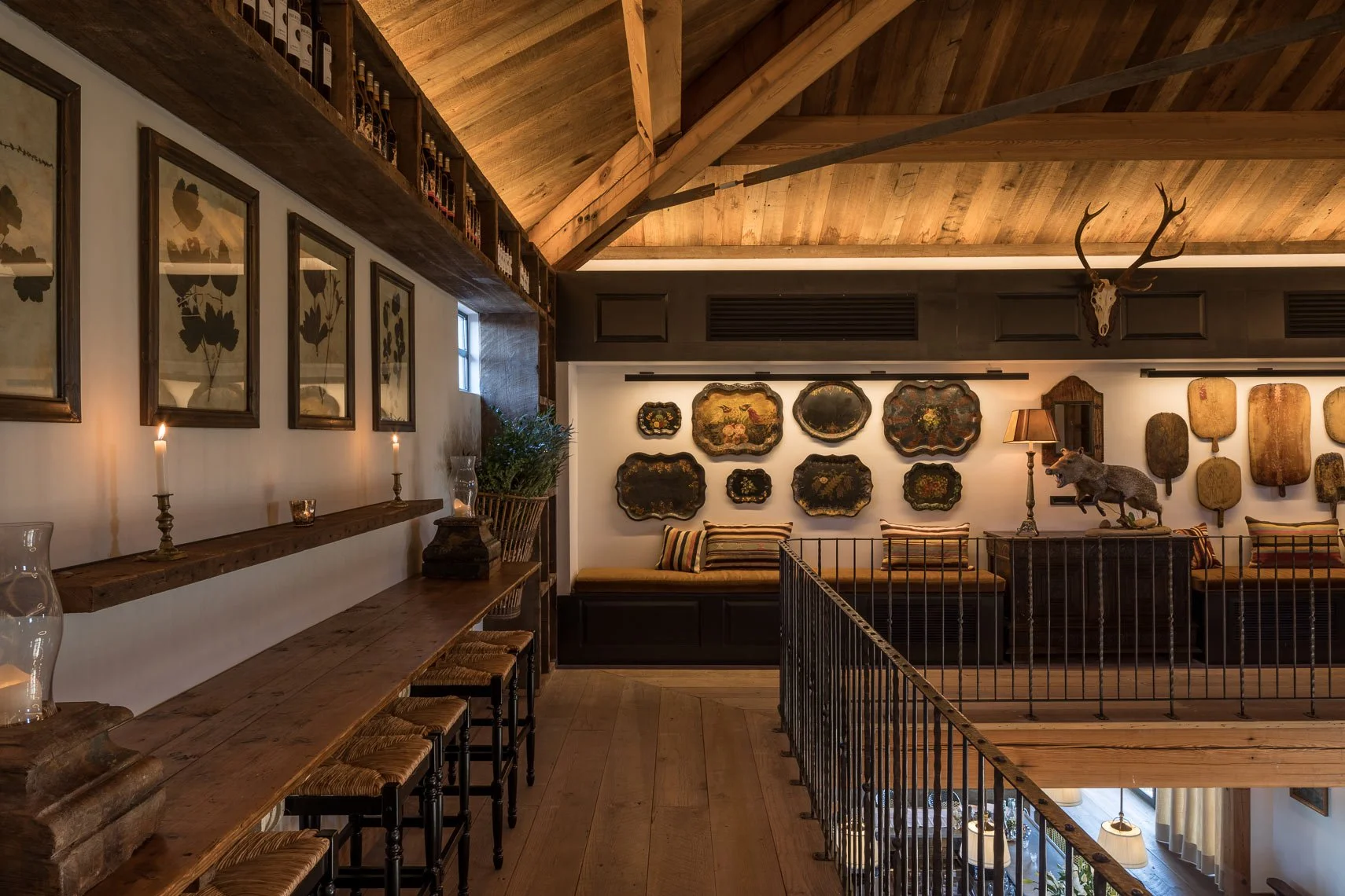A cozy living room with rustic wooden ceiling, framed botanical and decorative artwork on the walls, a wooden bench with striped cushions, a tall lamp, a decorative wolf figure, and a mounted deer skull with antlers.