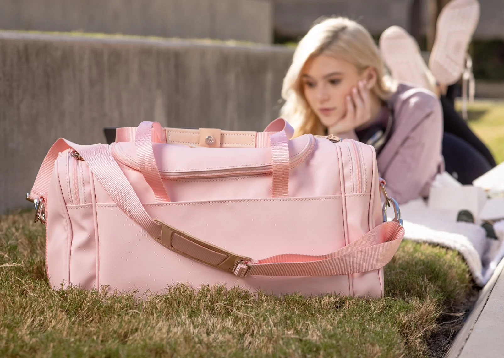 A pink duffel bag resting on the grass with a young woman lying on her stomach in the background, blurred, outdoors.