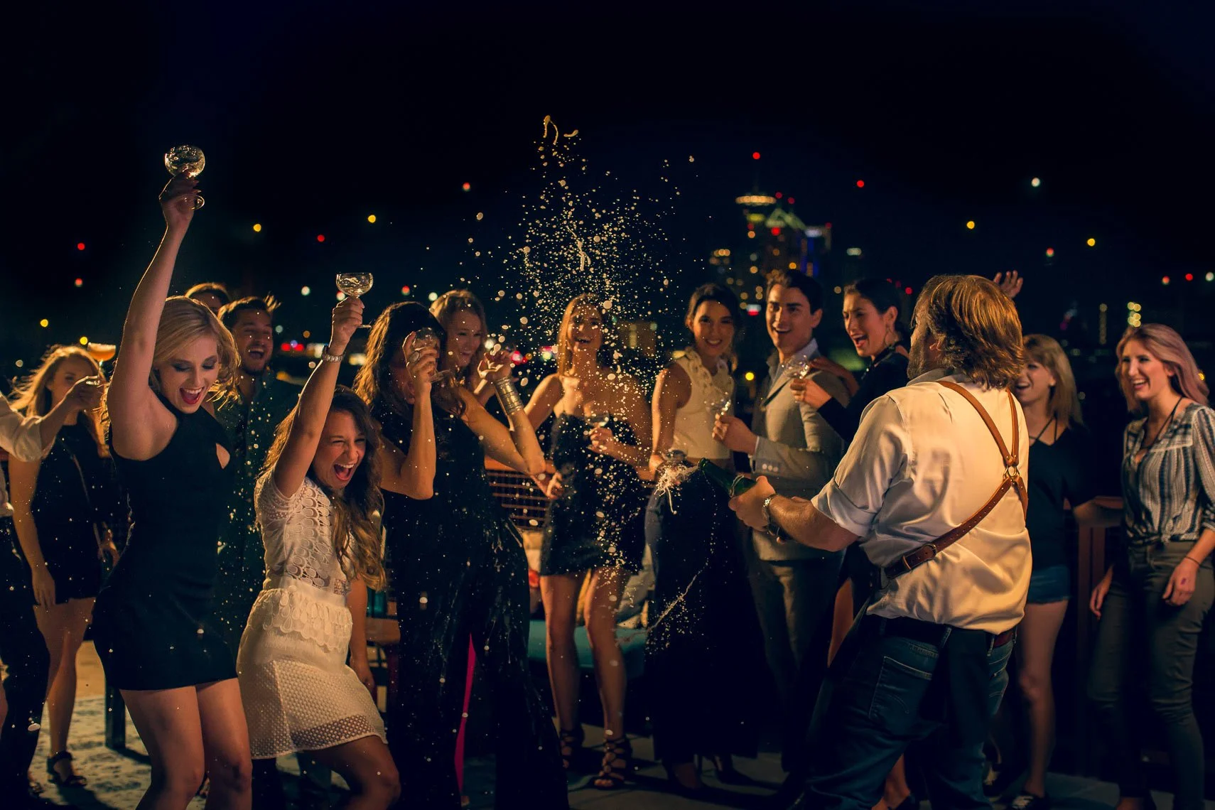 Group of people celebrating with champagne on a rooftop at night, party with joy, city skyline in the background.