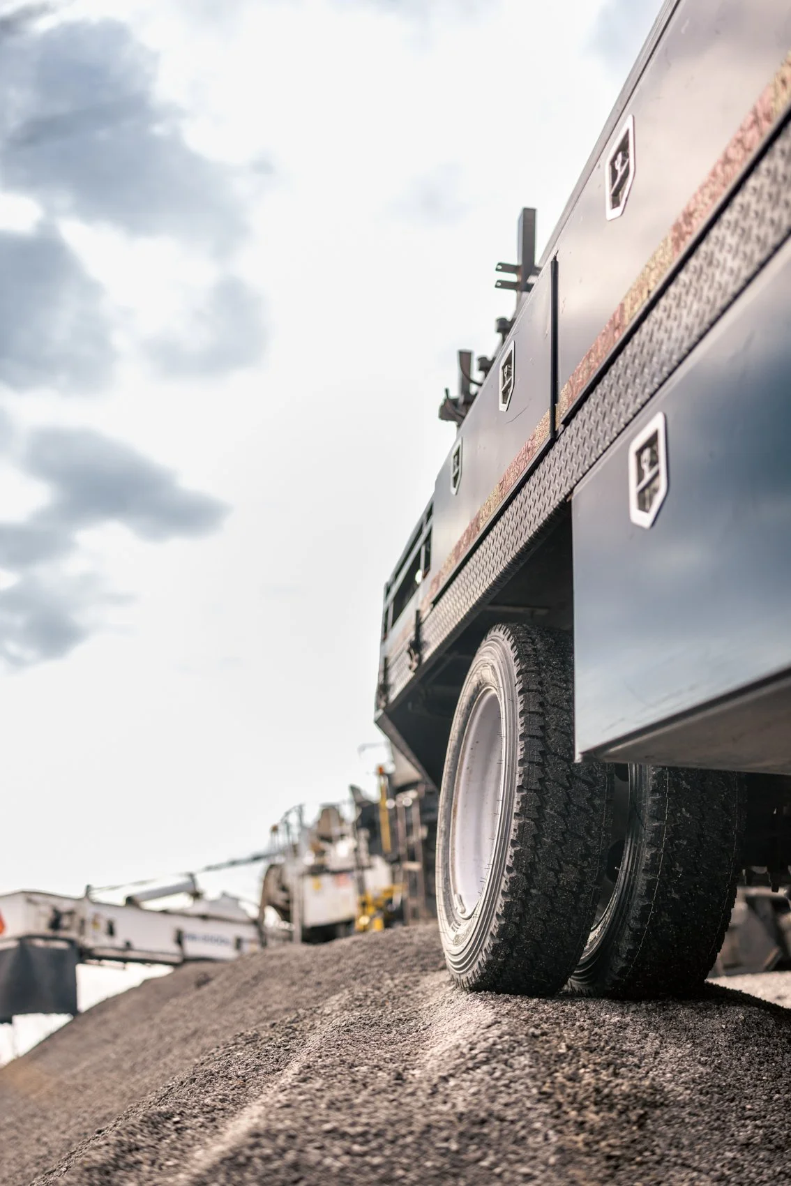 Close-up of a large truck tire on a gravel surface under a cloudy sky.