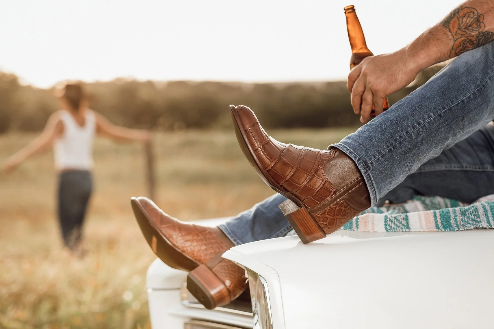Close-up of a man sitting on a white surface with his legs stretched out, wearing blue jeans and brown cowboy boots, holding a bottle of beer. In the background, a woman in a white top and dark pants is walking on a grassy field, with trees and a blu