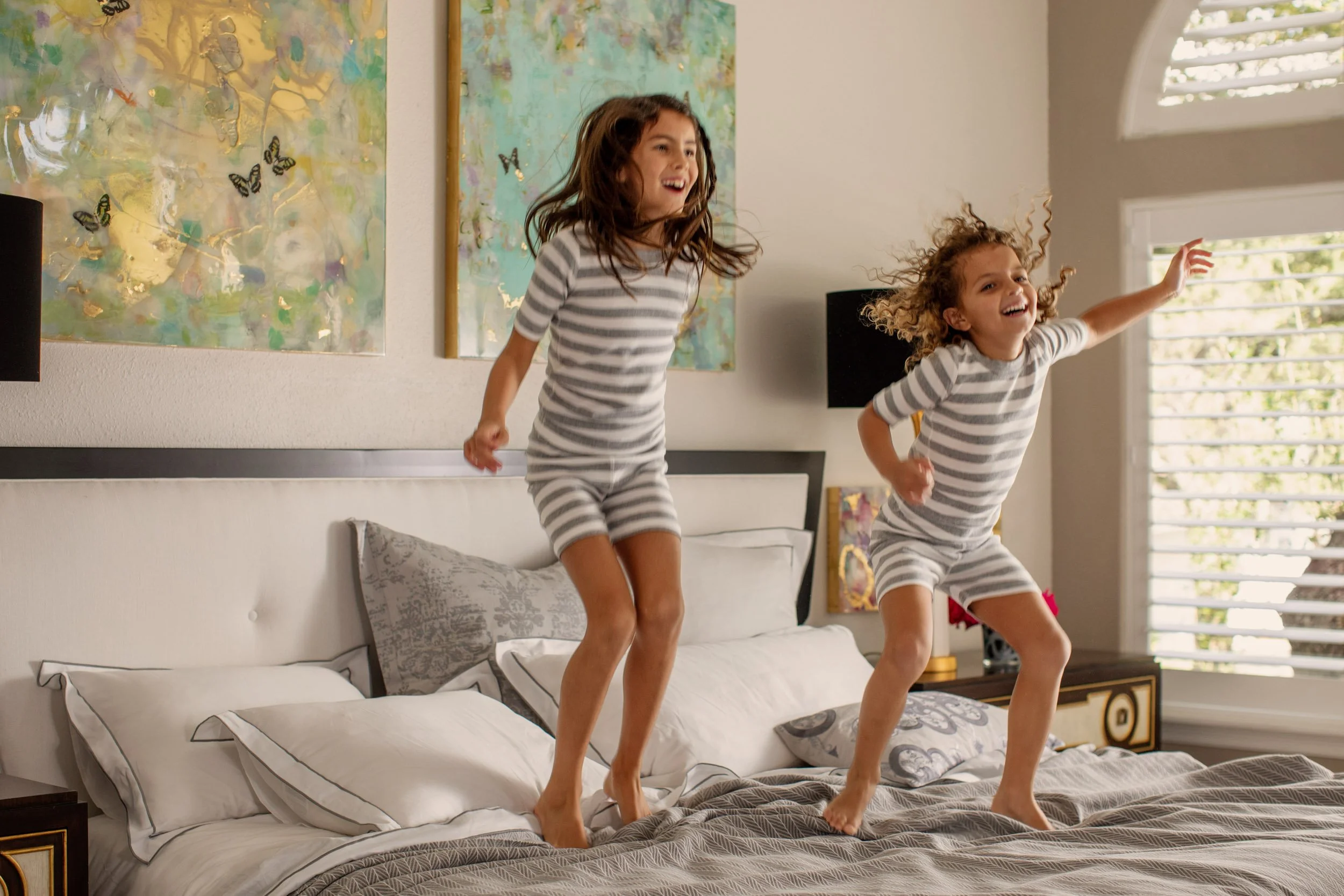 Two young girls jumping on a bed in a bright bedroom with abstract artwork and large windows, smiling and enjoying themselves.