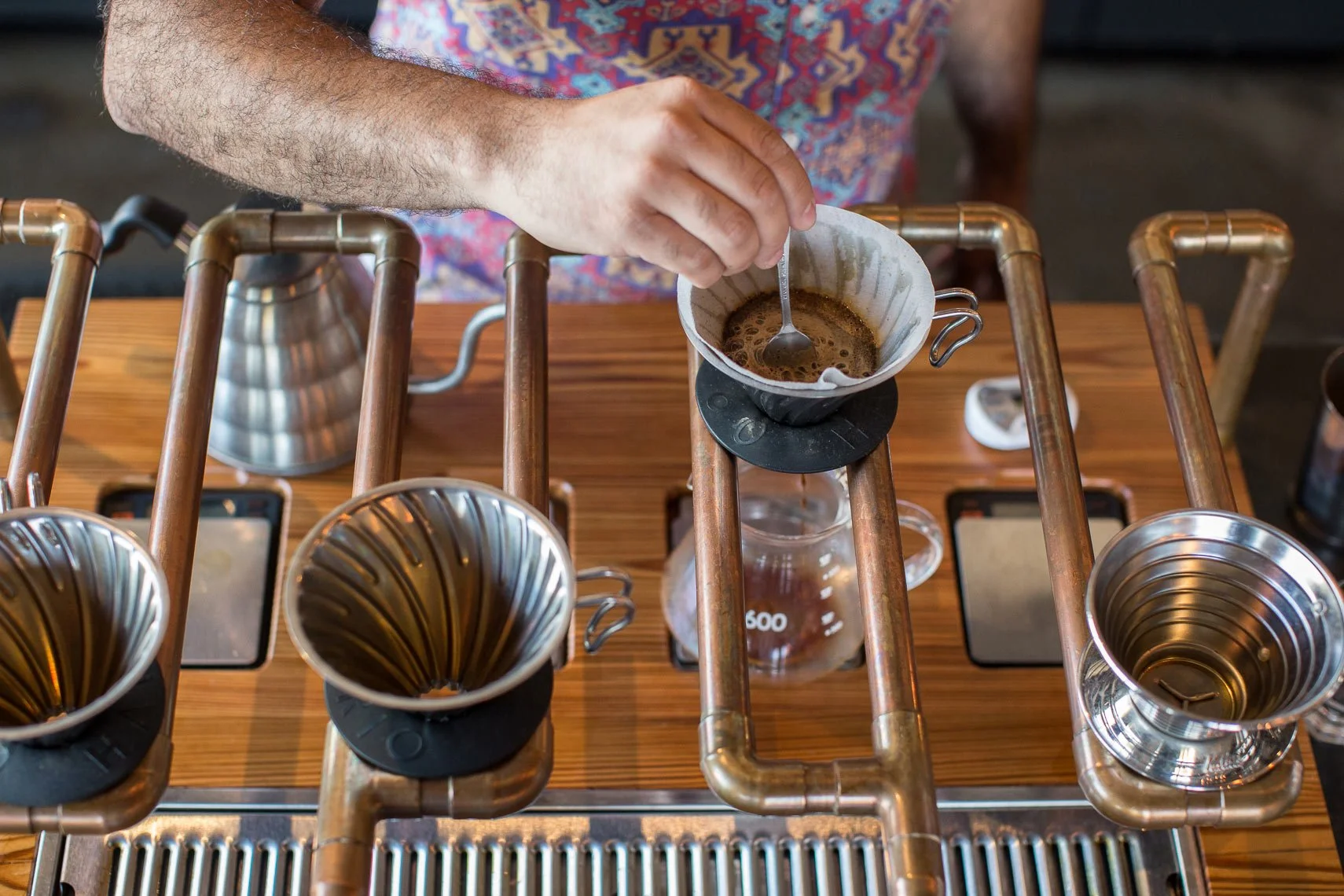 Overhead view of a person making pour-over coffee at a coffee shop with a copper pipe coffee stand, a metal gooseneck kettle, and empty coffee filters.