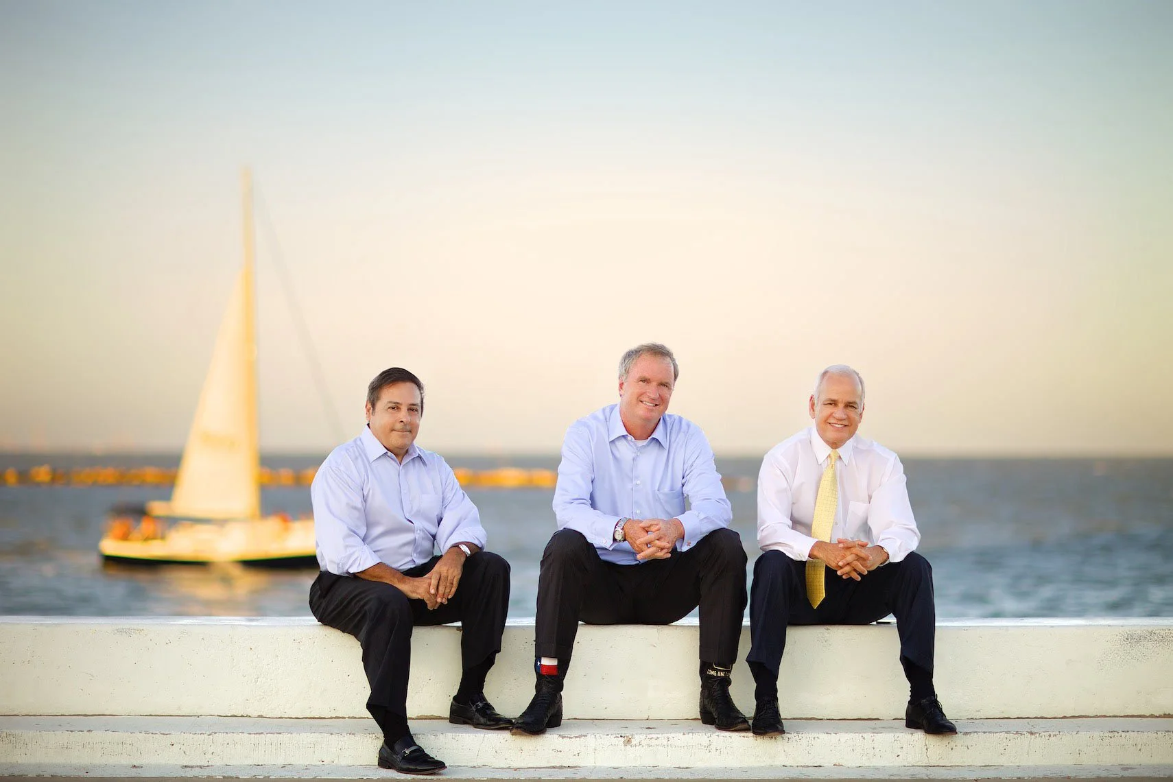Three men in business attire sitting on a ledge by the water with a sailboat in the background, during sunset.