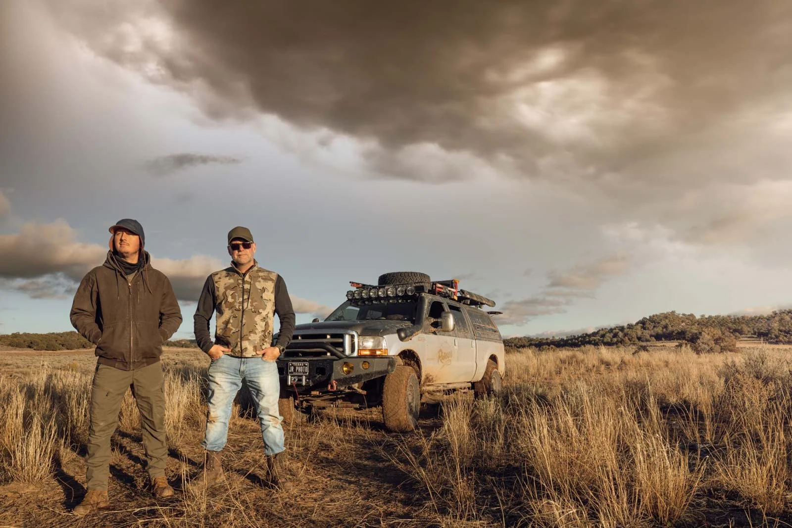 Two men stand in a grassy field with a rugged off-road vehicle behind them. The sky is cloudy and dramatic, suggesting an approaching storm.