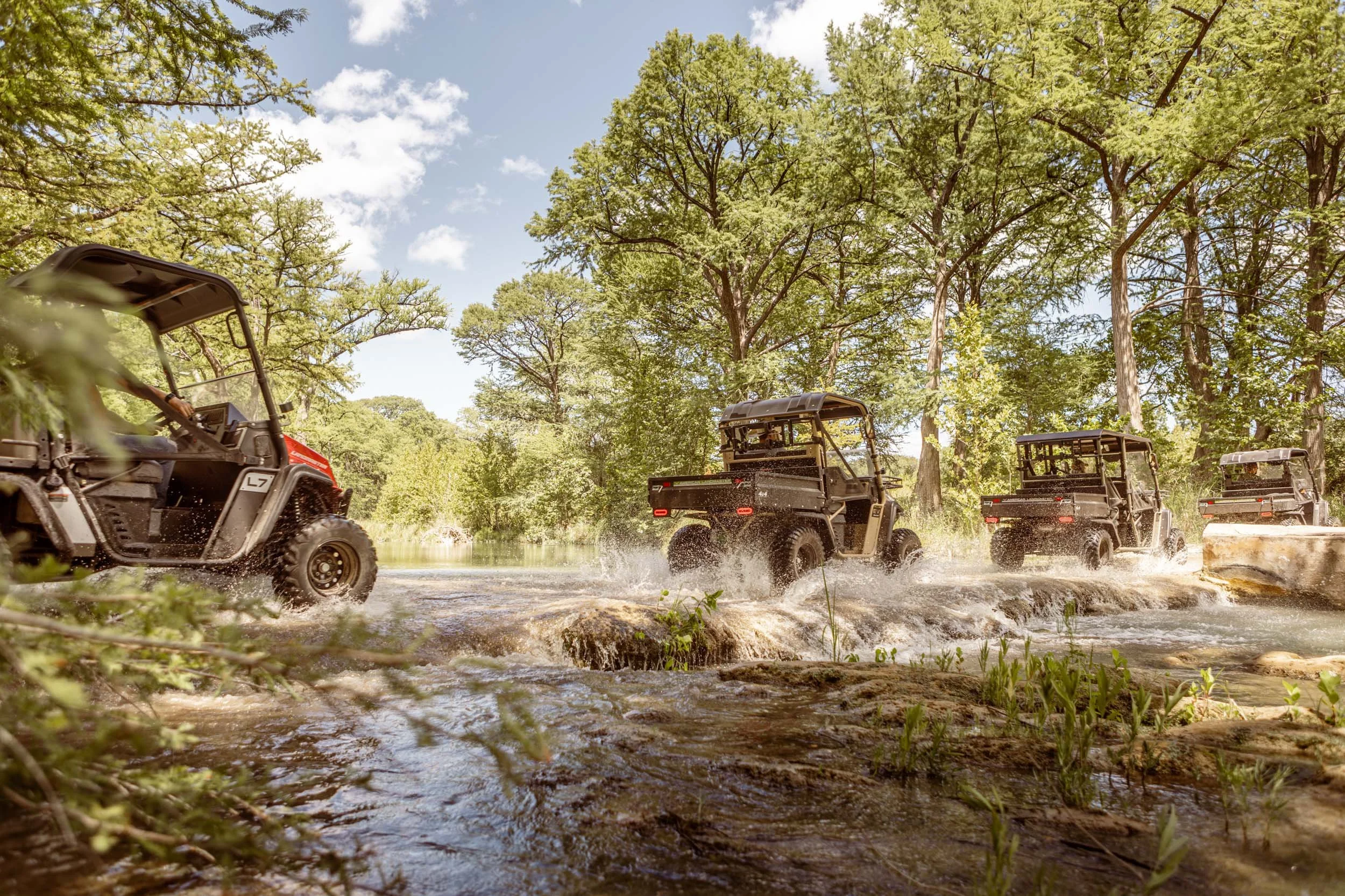 Several utility terrain vehicles driving through a shallow stream in a green forest under a partly cloudy sky.