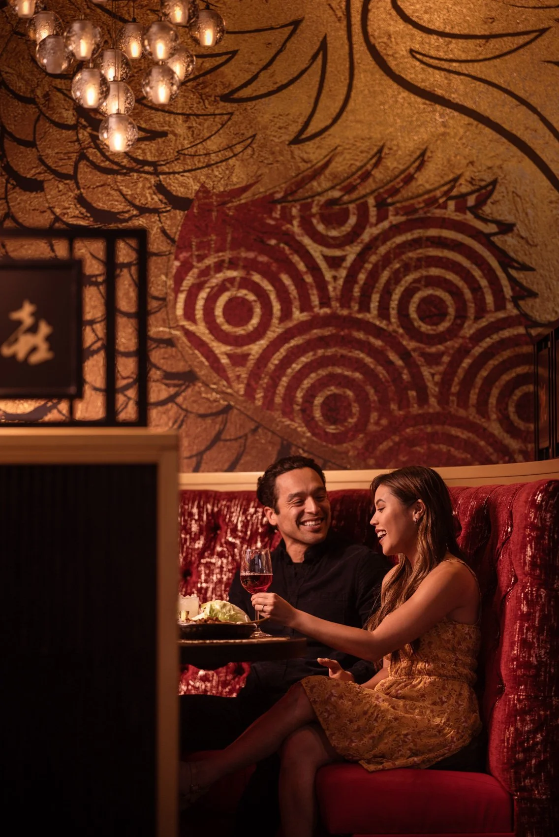 A man and woman sitting on a red velvet booth in a dimly lit restaurant, smiling and laughing while sharing a drink of red wine. The background features an artistic wall with abstract patterns and a chandelier hanging above.
