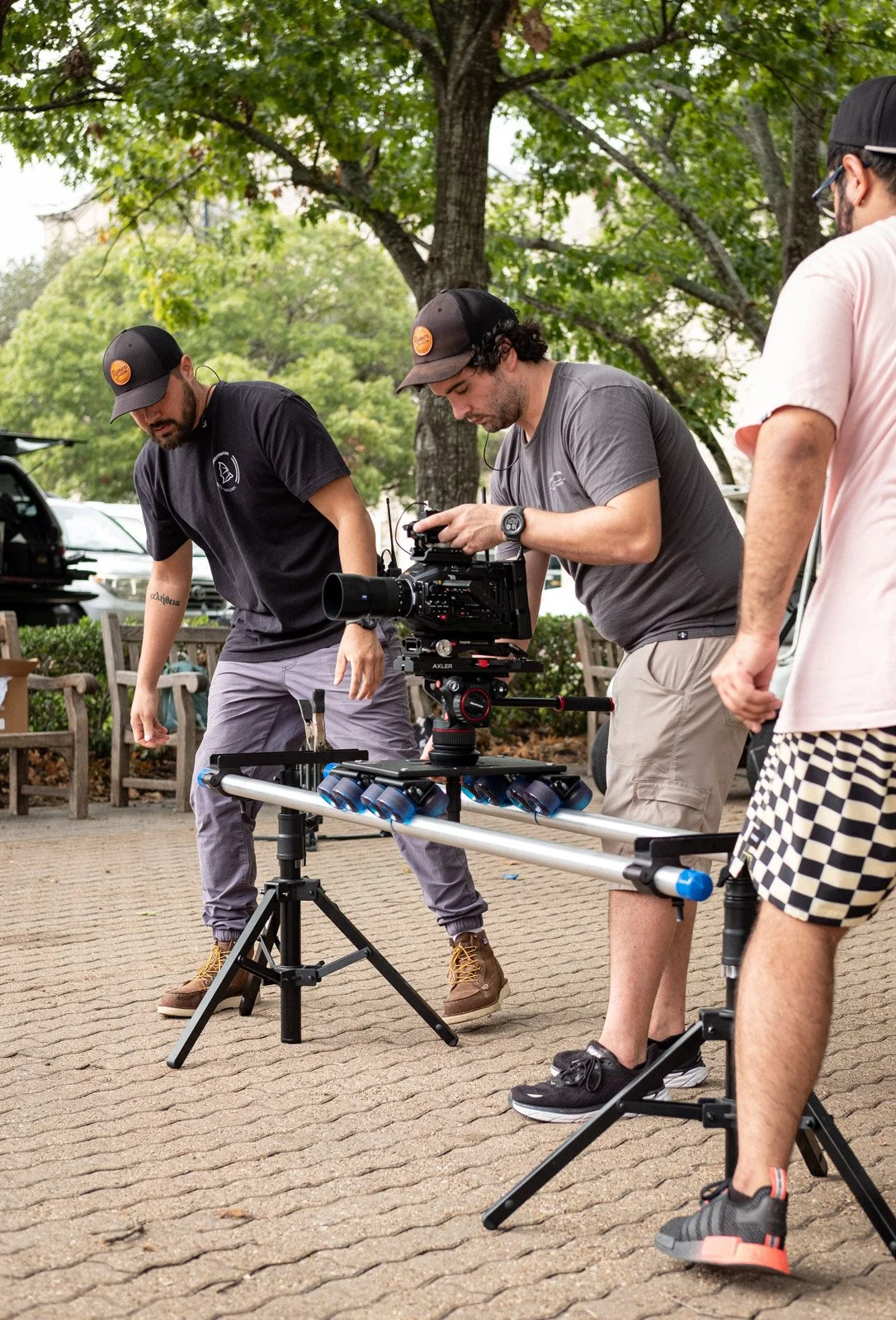 A group of three men working with camera equipment outdoors on a cloudy day, set up on a tripod on a paved area with trees and parked cars in the background.