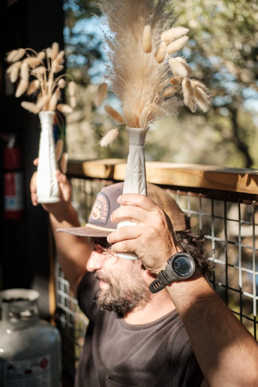 A man wearing a hat and a watch is holding a white vase with dried grasses, smiling in an outdoor setting.