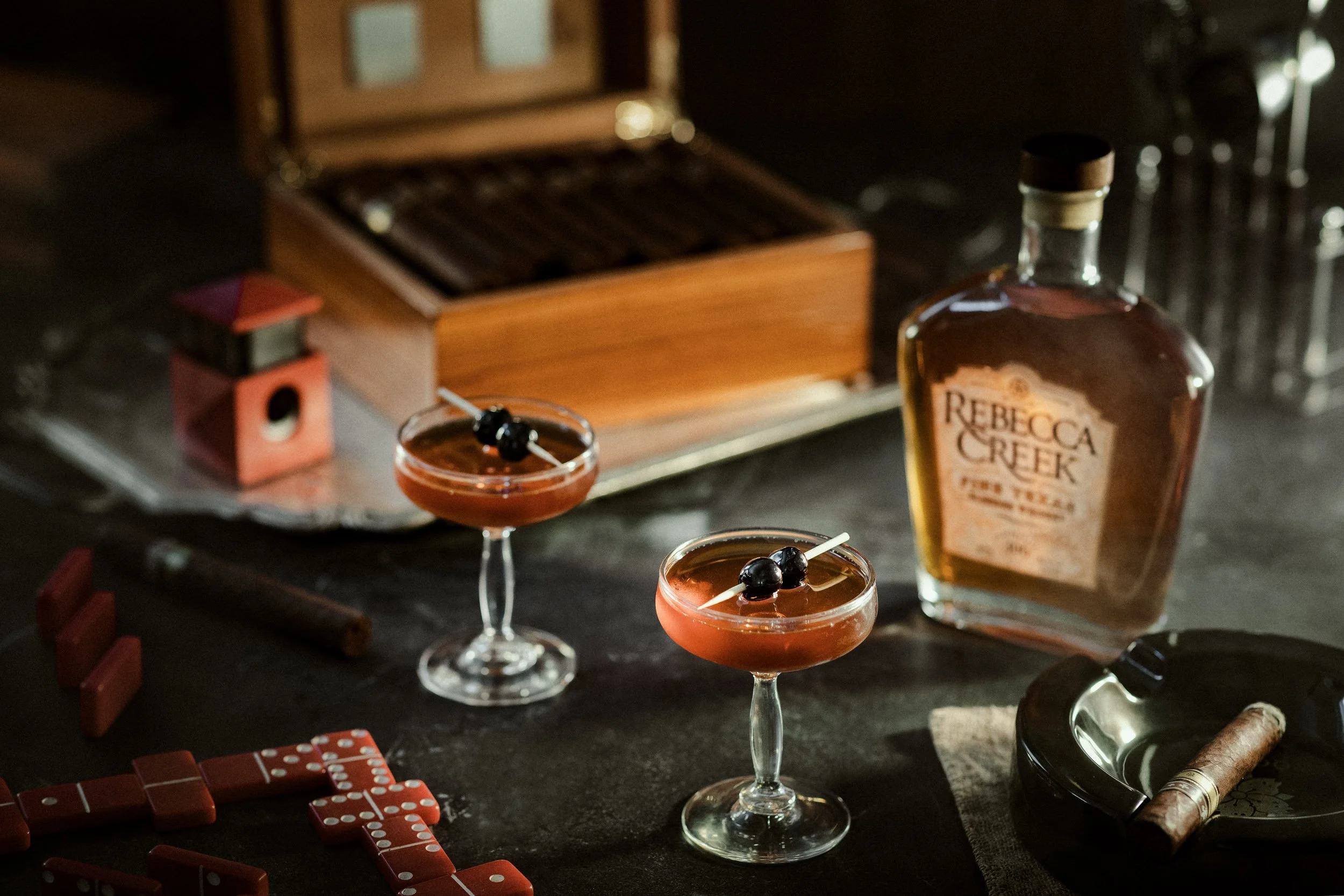 Two cocktail glasses filled with a dark red drink, garnished with cherries on toothpicks, set on a dark surface alongside a bottle labeled 'Rebecca Creek' whiskey, a cigar, and scattered red domino tiles.