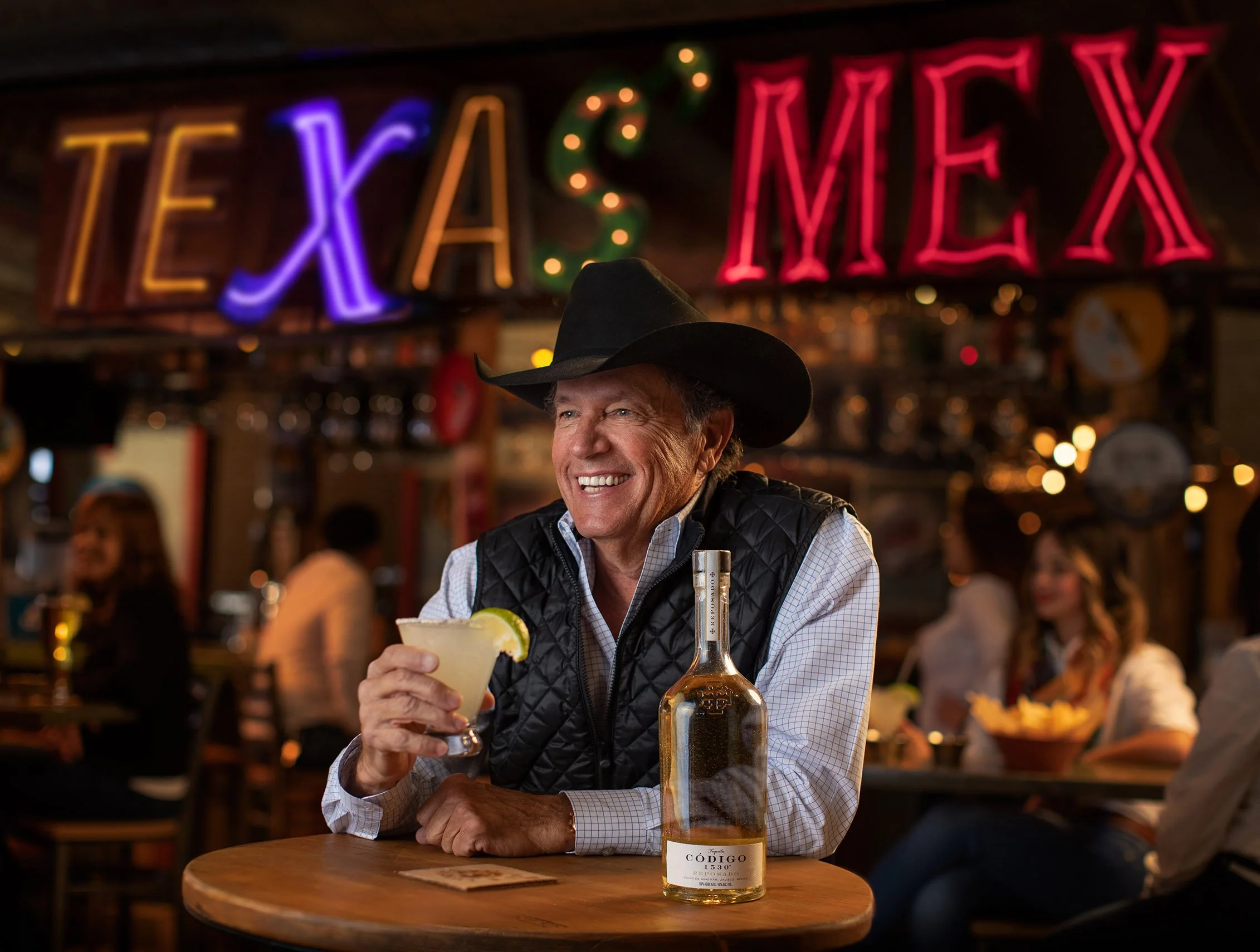 A smiling man wearing a black cowboy hat and a black quilted vest sitting at a wooden table in a bar or restaurant, holding a margarita glass with a lime wedge, with a bottle of Código tequila on the table in front, and a neon sign that says 'TEXAS' 