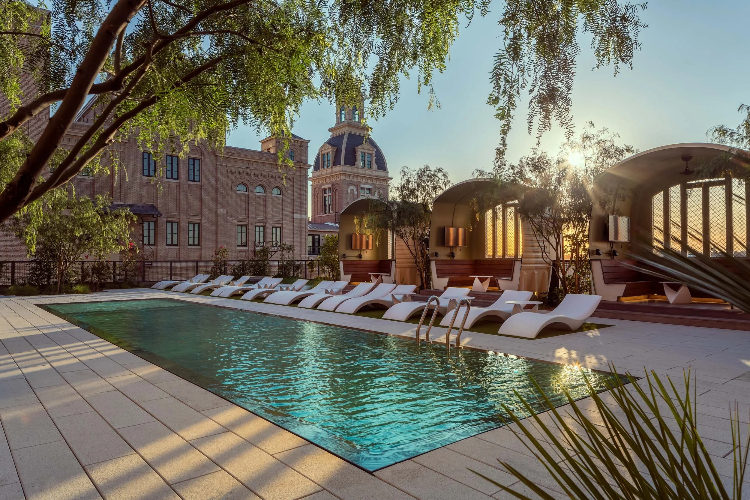 A pool area with lounge chairs, shaded cabanas, and surrounded by trees, with a historic building with clock tower in the background and the sun setting.