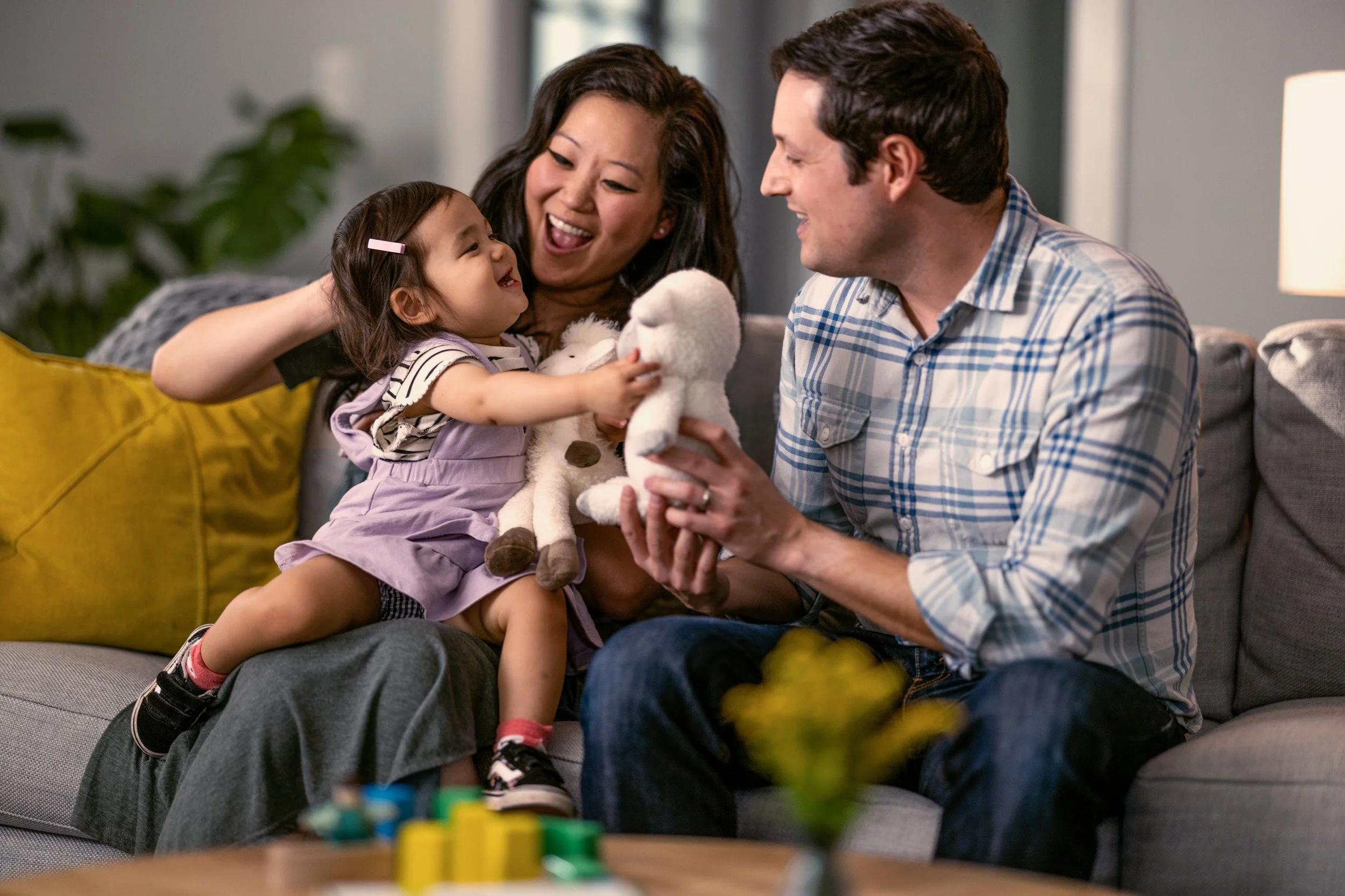 A family of three sitting on a gray sofa in a living room, smiling and interacting with each other. The young girl, holding a stuffed animal, is laughing while looking at the adult woman and man, who are also smiling.