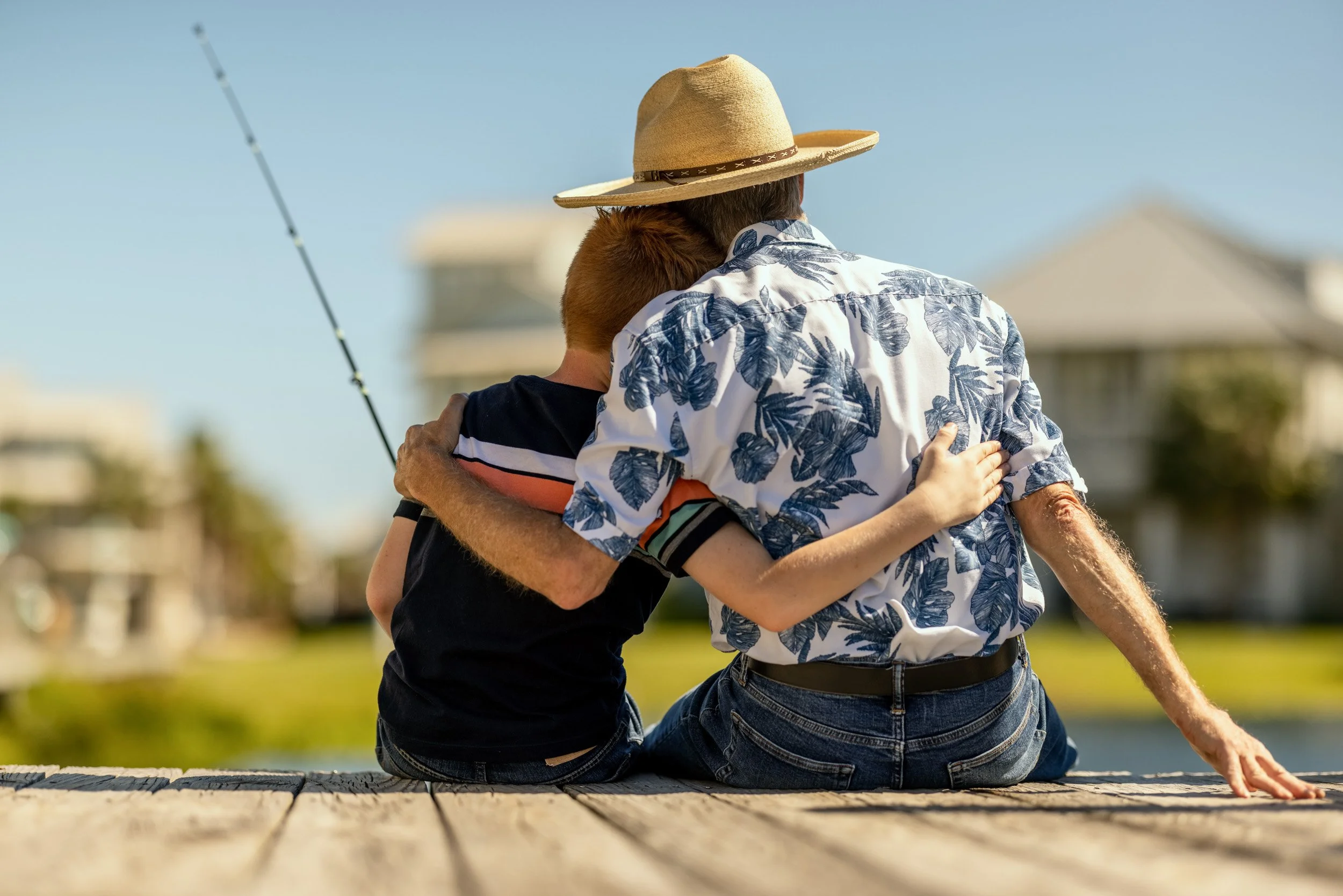 Two people hugging each other while sitting on a wooden dock outdoors, with a lake and buildings in the background, under clear blue skies.