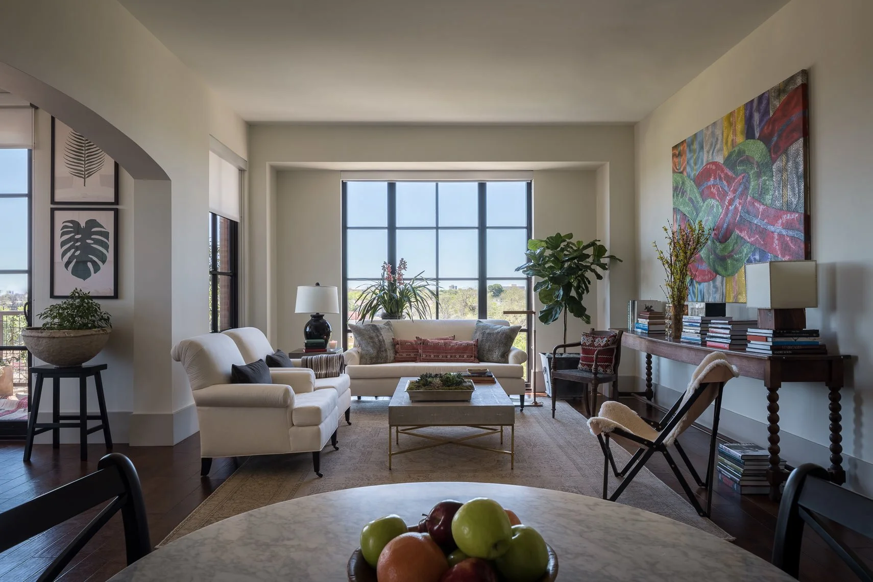 A living room with white sofas, a stone coffee table, and plants near large windows, decorated with artwork and books.