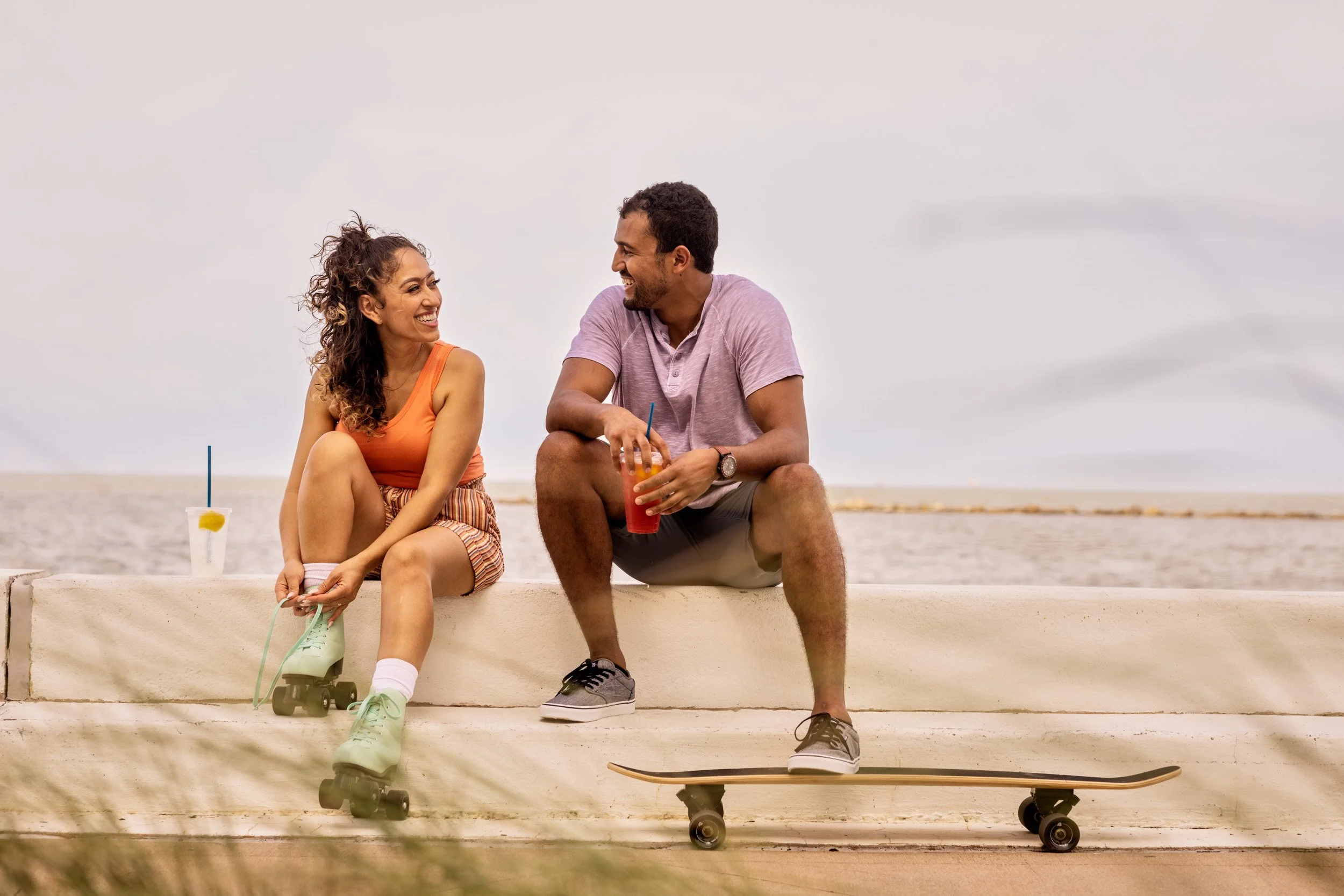 A woman and a man sitting on a ledge at the beach. The woman is wearing a sleeveless orange top and striped shorts, with her roller skates and a drink with a straw beside her. The man is wearing a light purple T-shirt, shorts, and sneakers, holding a