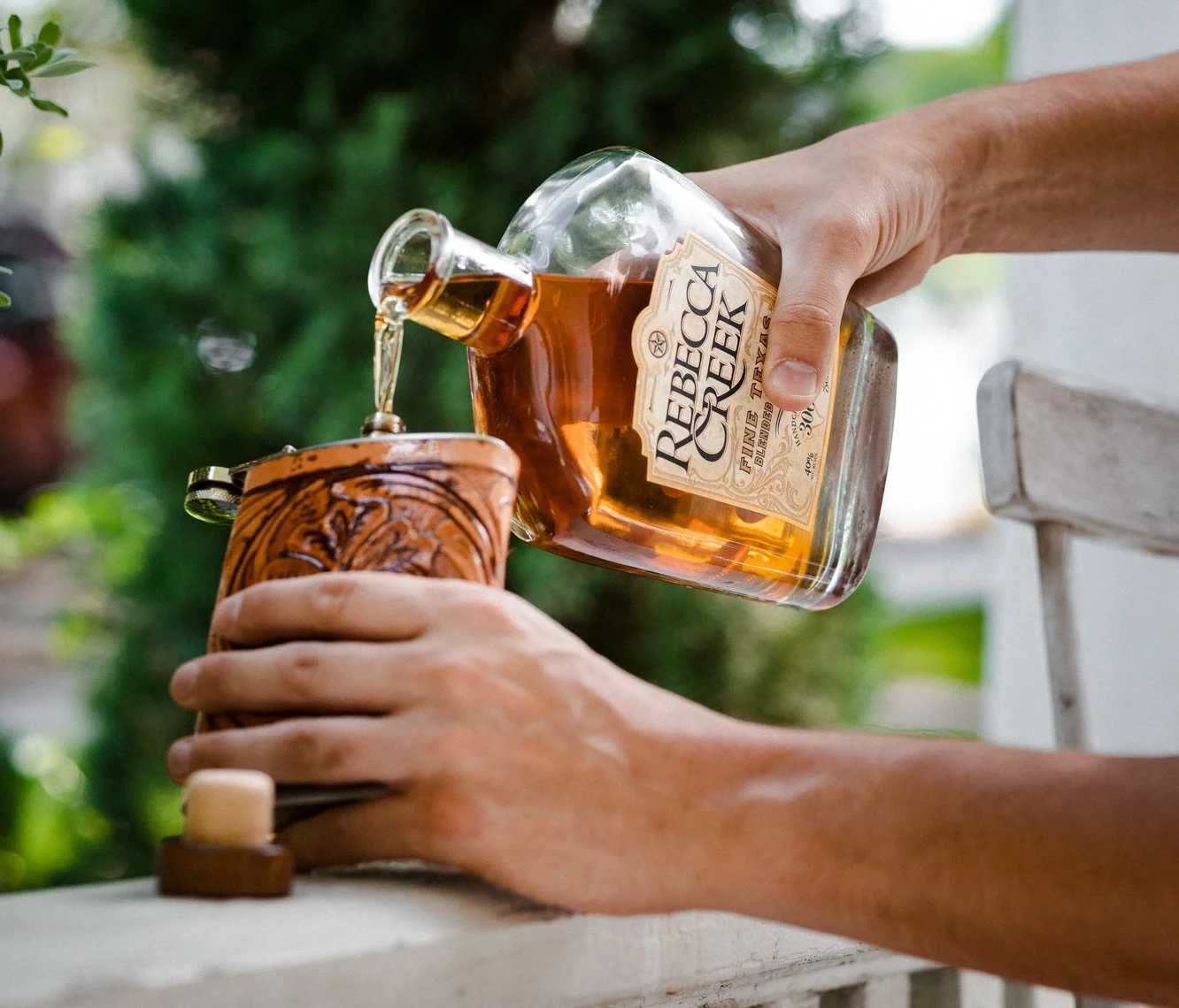Person pouring whiskey from a bottle labeled 'Rebeca Creek' into a glass held with the other hand, outdoors with green foliage in the background.