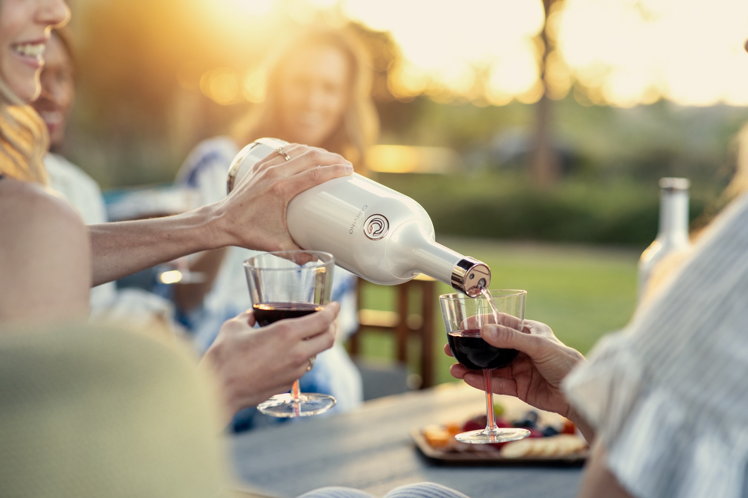 People sharing a glass of red wine during an outdoor gathering at sunset, with a woman pouring wine.