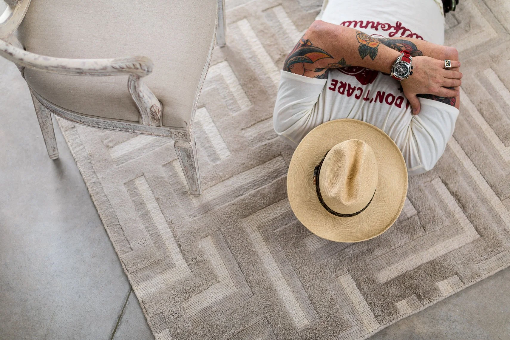 From above view of a person with tattoos on their arms sitting on a beige rug with a cowboy hat on their lap, wearing a white t-shirt with red lettering and a red watch. There is a vintage-style chair with a distressed white paint finish and light-co
