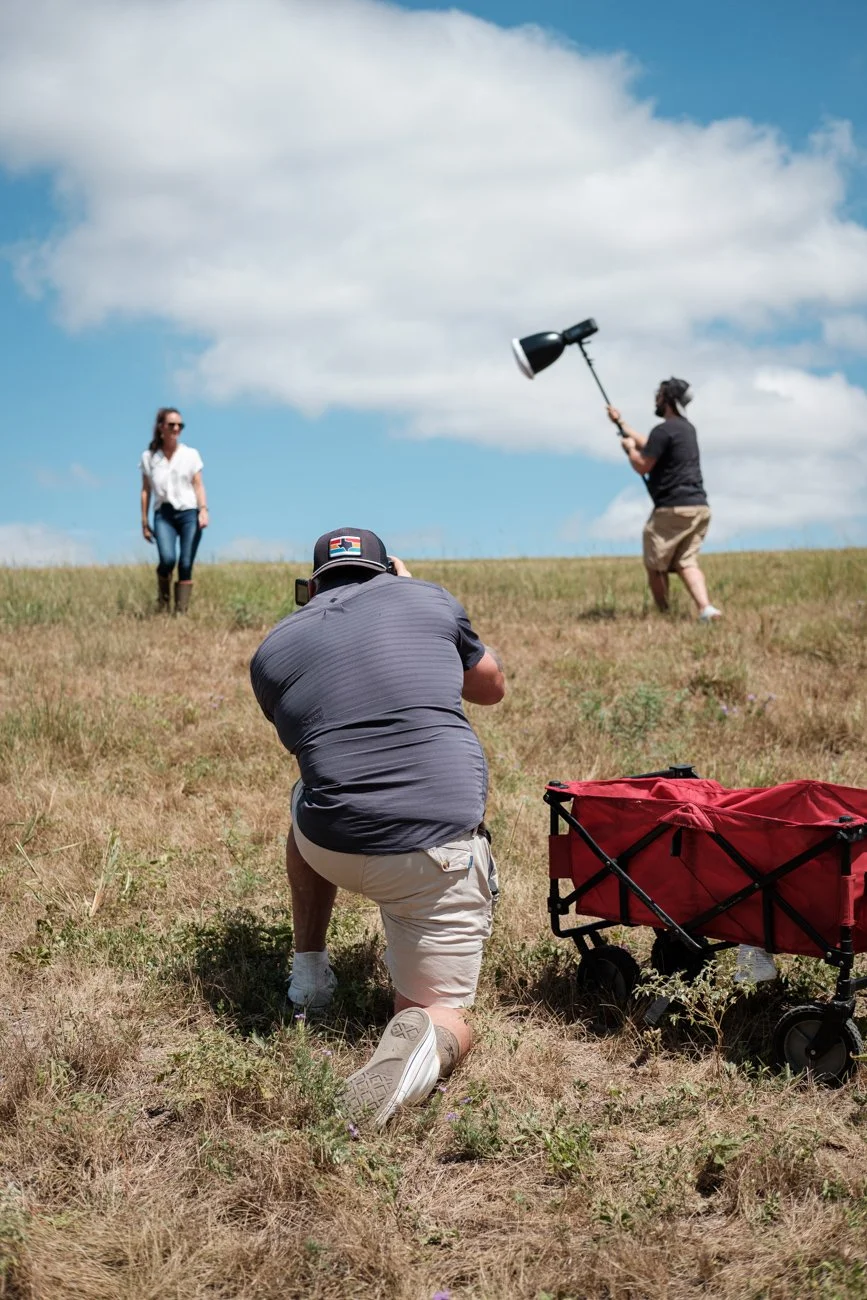 A woman standing on a grassy hill, a man kneeling with a camera, and another person holding a large spotlight against a partly cloudy sky.