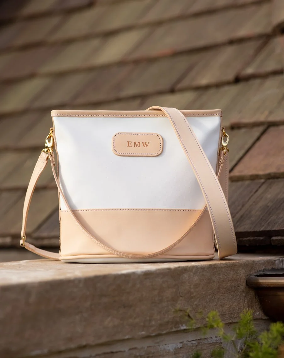 A beige and white handbag with a pink leather tag branded 'EMW,' resting on a wooden surface with a wooden shingle background and some green plants at the bottom.