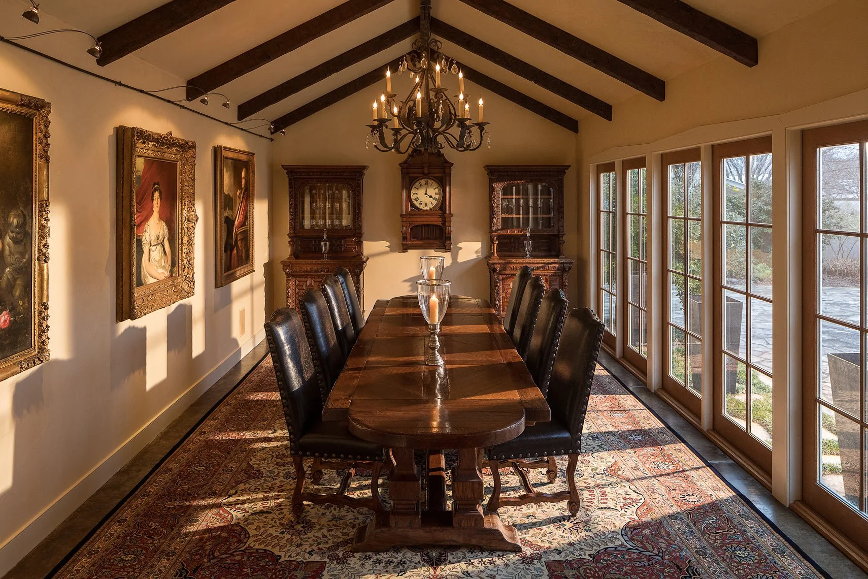 Elegant dining room with a long wooden table, high-back upholstered chairs, candles in glass holders, large windows on the right, dark wood beams on the ceiling, antique paintings on the left wall, an ornate clock and cabinets at the far end, and a c