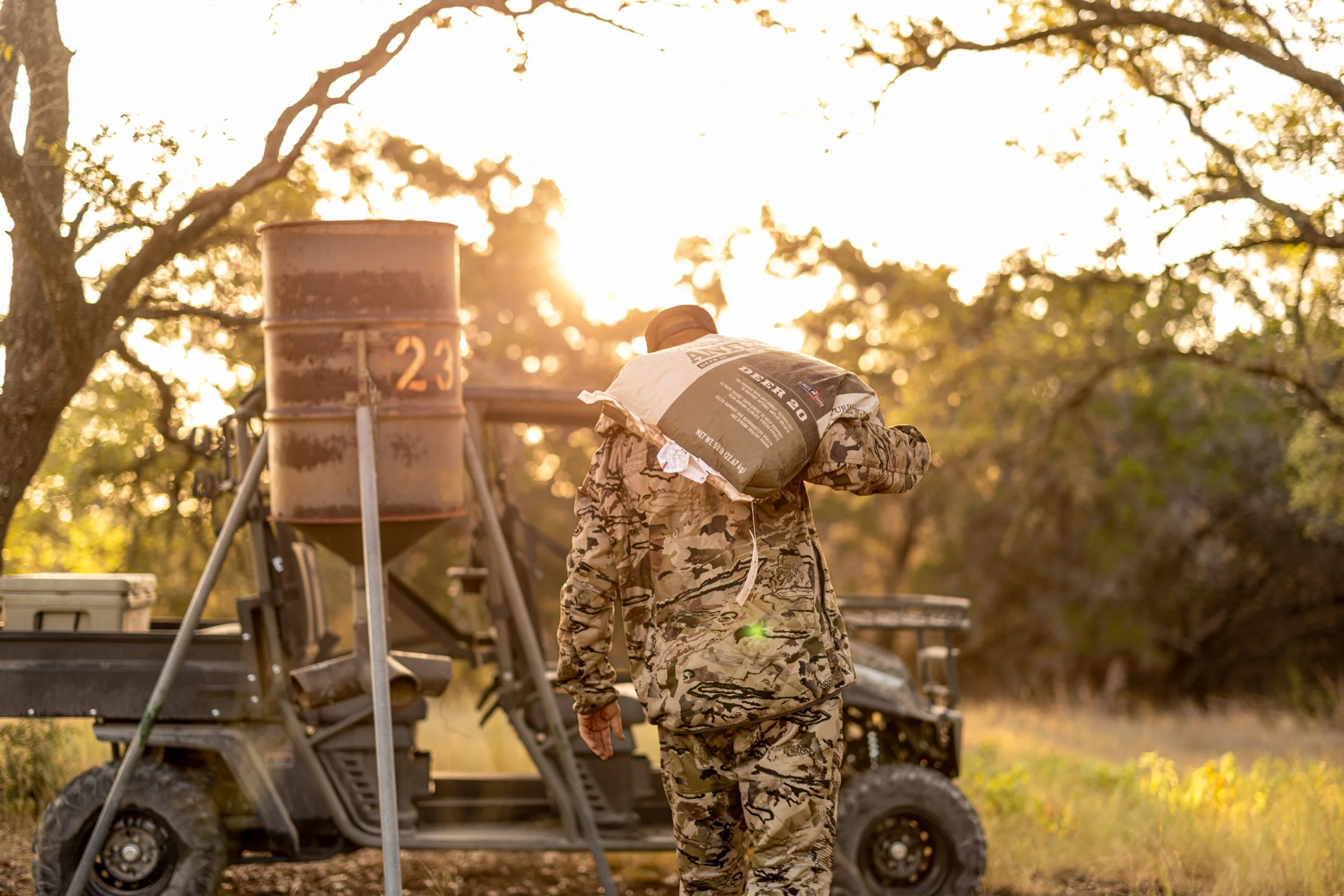 A person dressed in camouflage military uniform carrying a bag, standing outdoors near equipment and a metal barrel, during sunset in a wooded area.