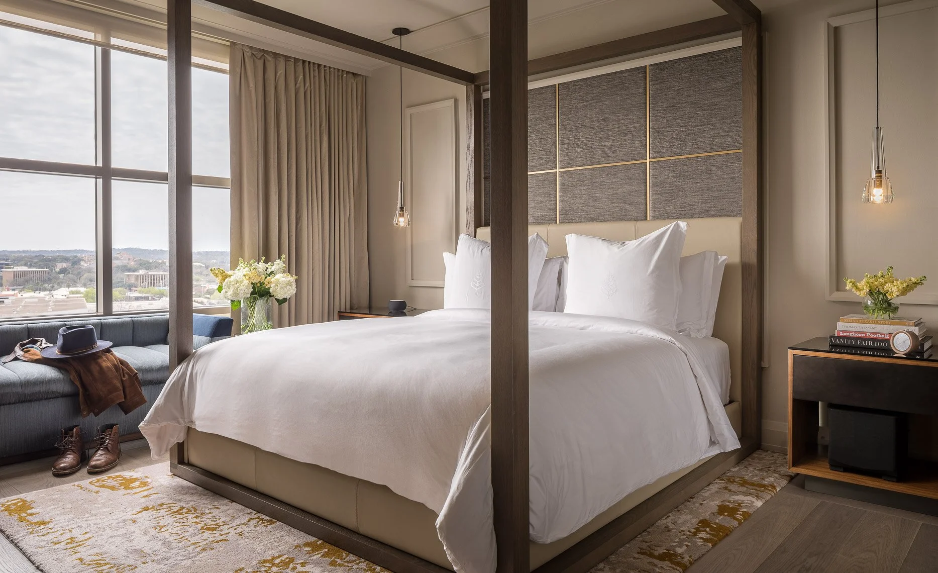 Modern hotel bedroom with a large window, bed with white linens, nightstands with flowers and books, and a seating area by the window with a hat, jacket, and boots.
