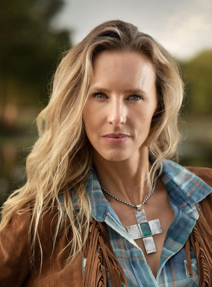 Close-up of a woman with wavy blonde hair, wearing a blue plaid shirt, brown fringed jacket, and a cross-shaped silver necklace. She has a serious expression and is outdoors with blurred trees in the background.