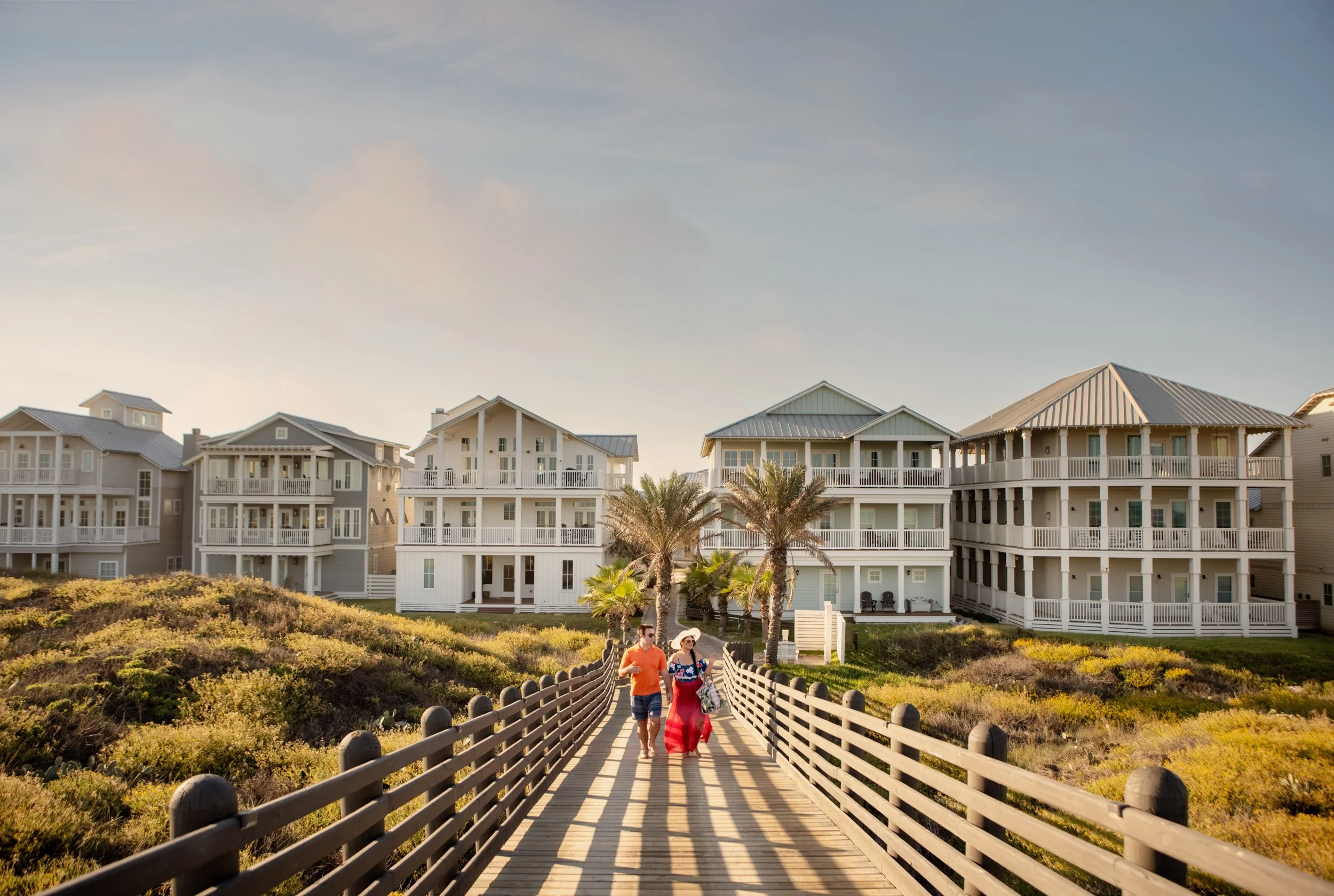 Two women walking on a wooden boardwalk toward white apartment buildings with a mostly clear sky above, with some clouds and sunlight.