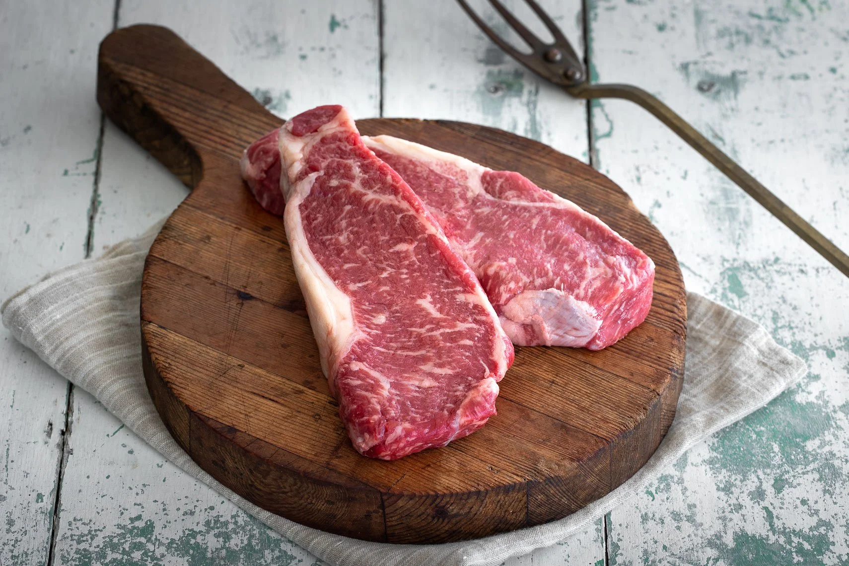 Two raw ribeye steaks on a round wooden cutting board on a white distressed wooden surface with a white cloth underneath.