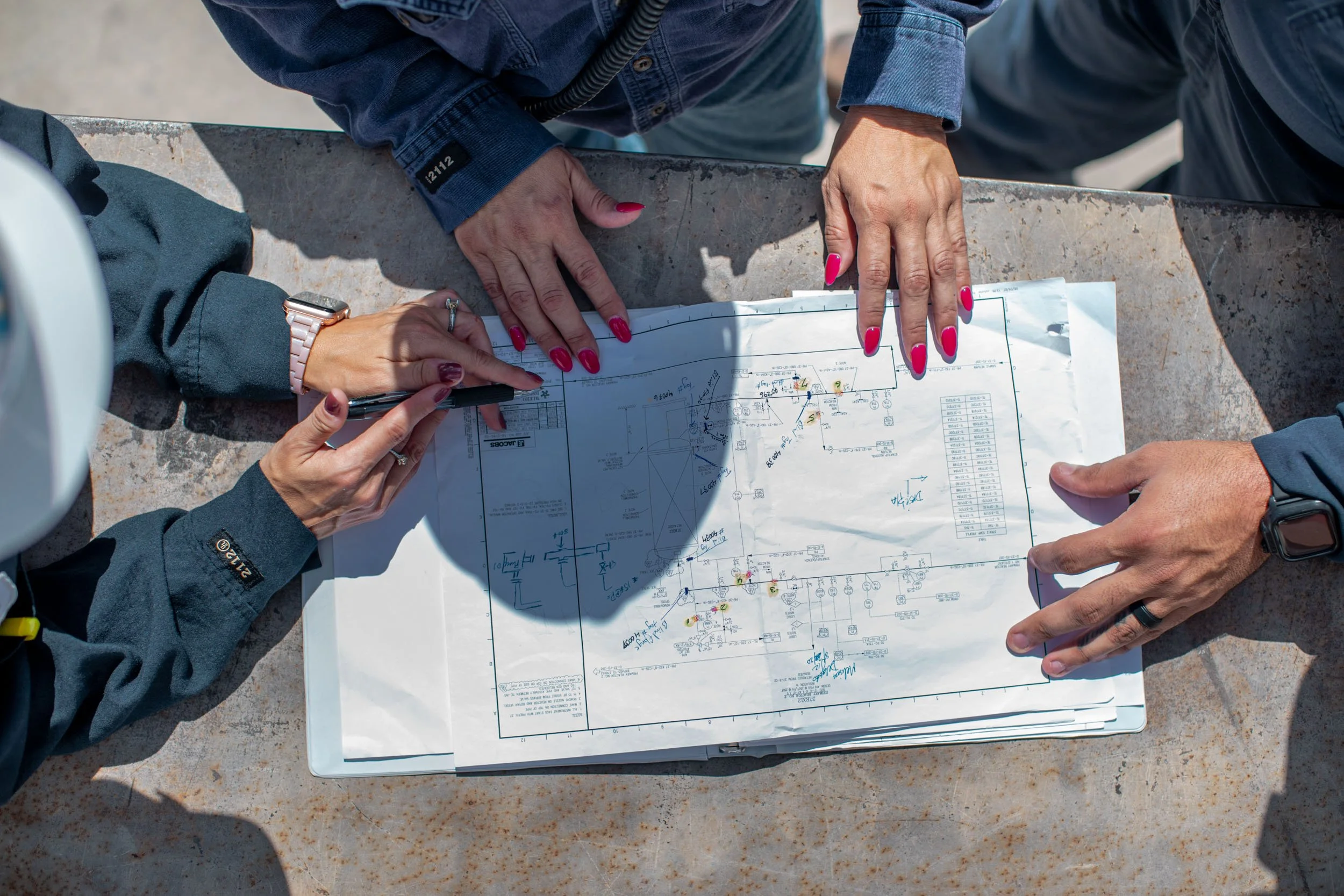 Two people review architectural or engineering plans outdoors on a stone surface, with detailed blueprints and markings.